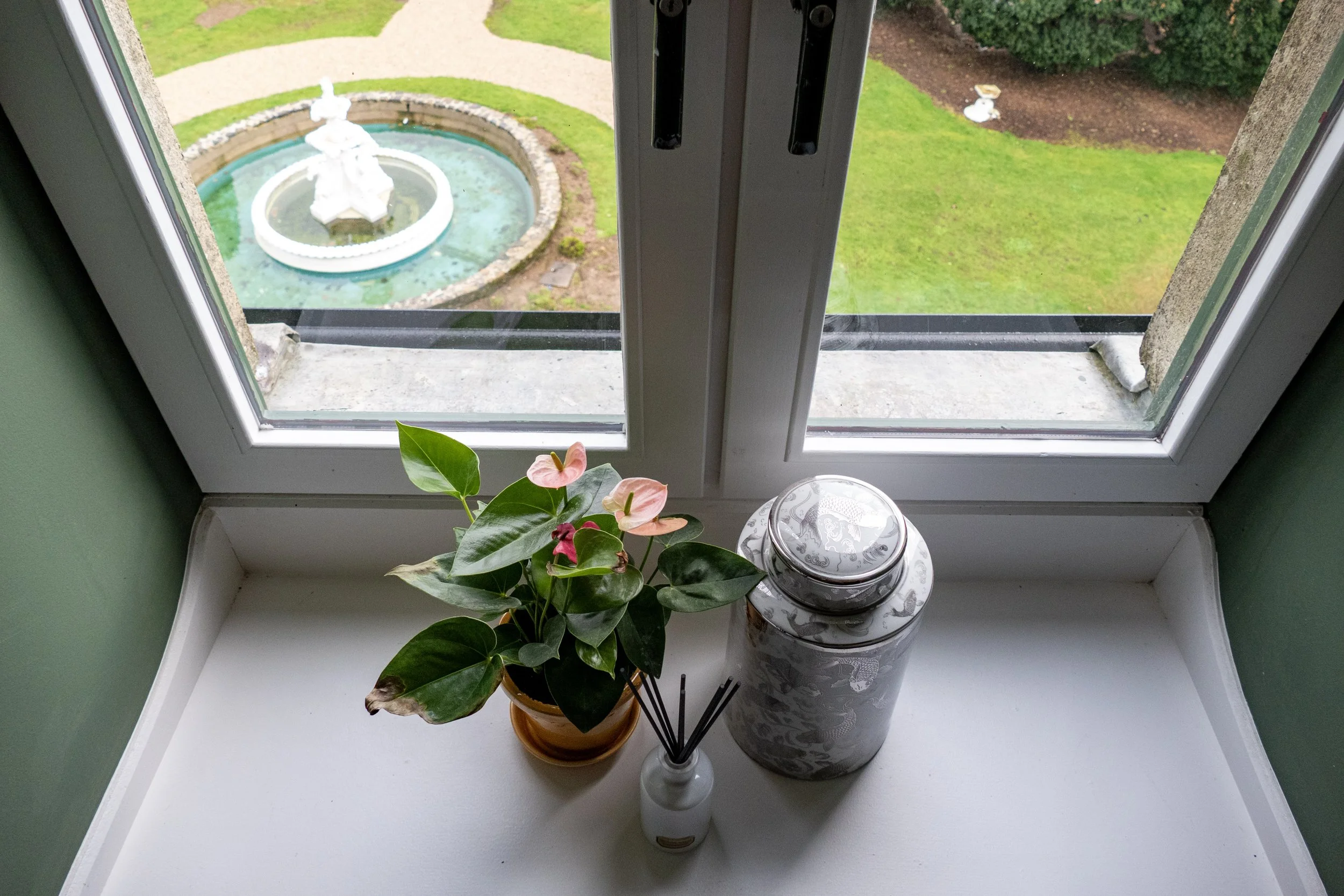 View through a window showing a garden with a fountain and green grass; on the windowsill, a potted plant with pink and green leaves, a white reed diffuser, and a decorative silver jar.