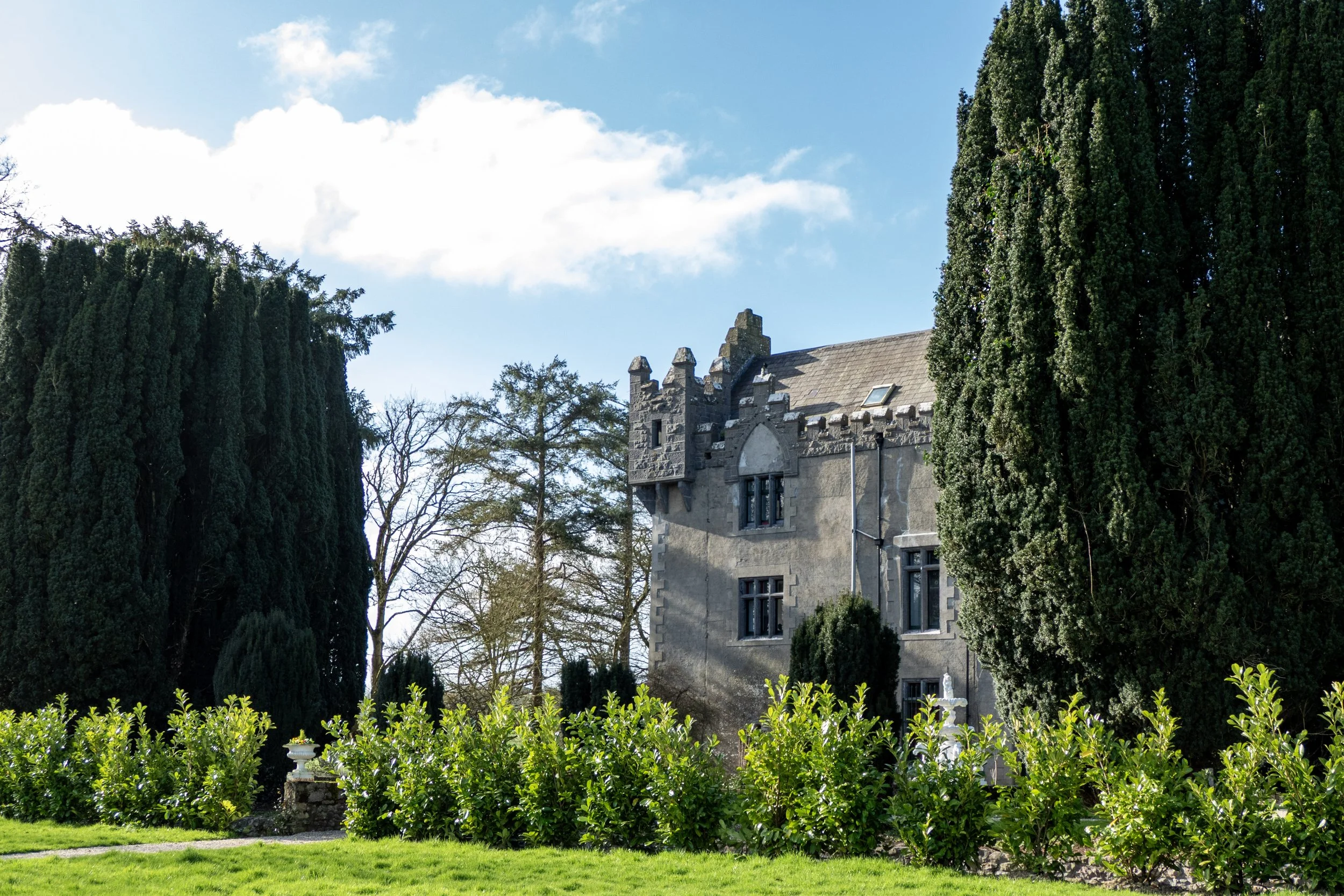 A large stone house with a castle-like turret, surrounded by tall evergreen trees and a well-maintained green garden on a sunny day.