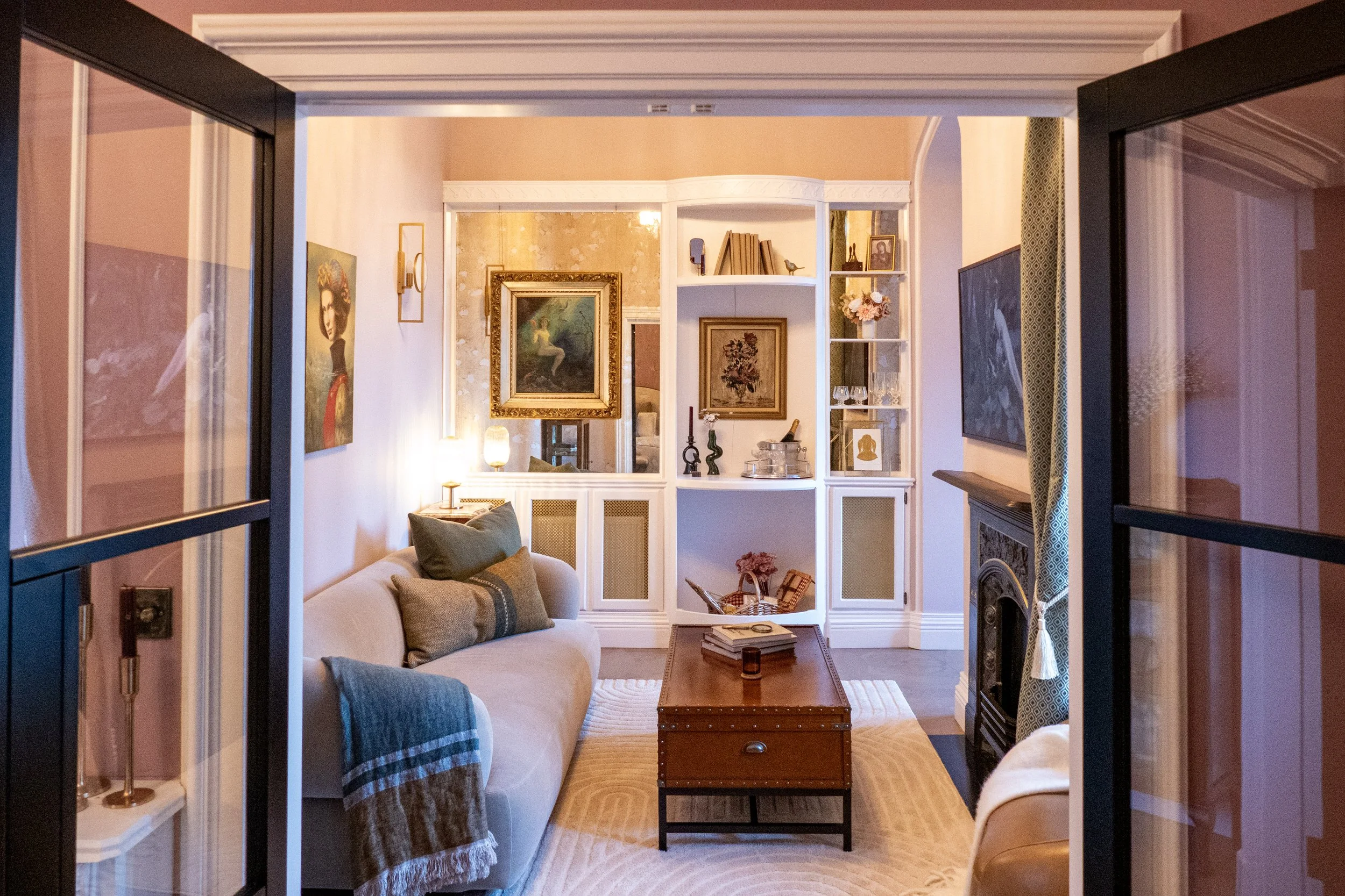 Cozy living room with two sofas, a wooden coffee table, built-in bookshelf, and artwork on the walls, seen through open black framed glass doors.