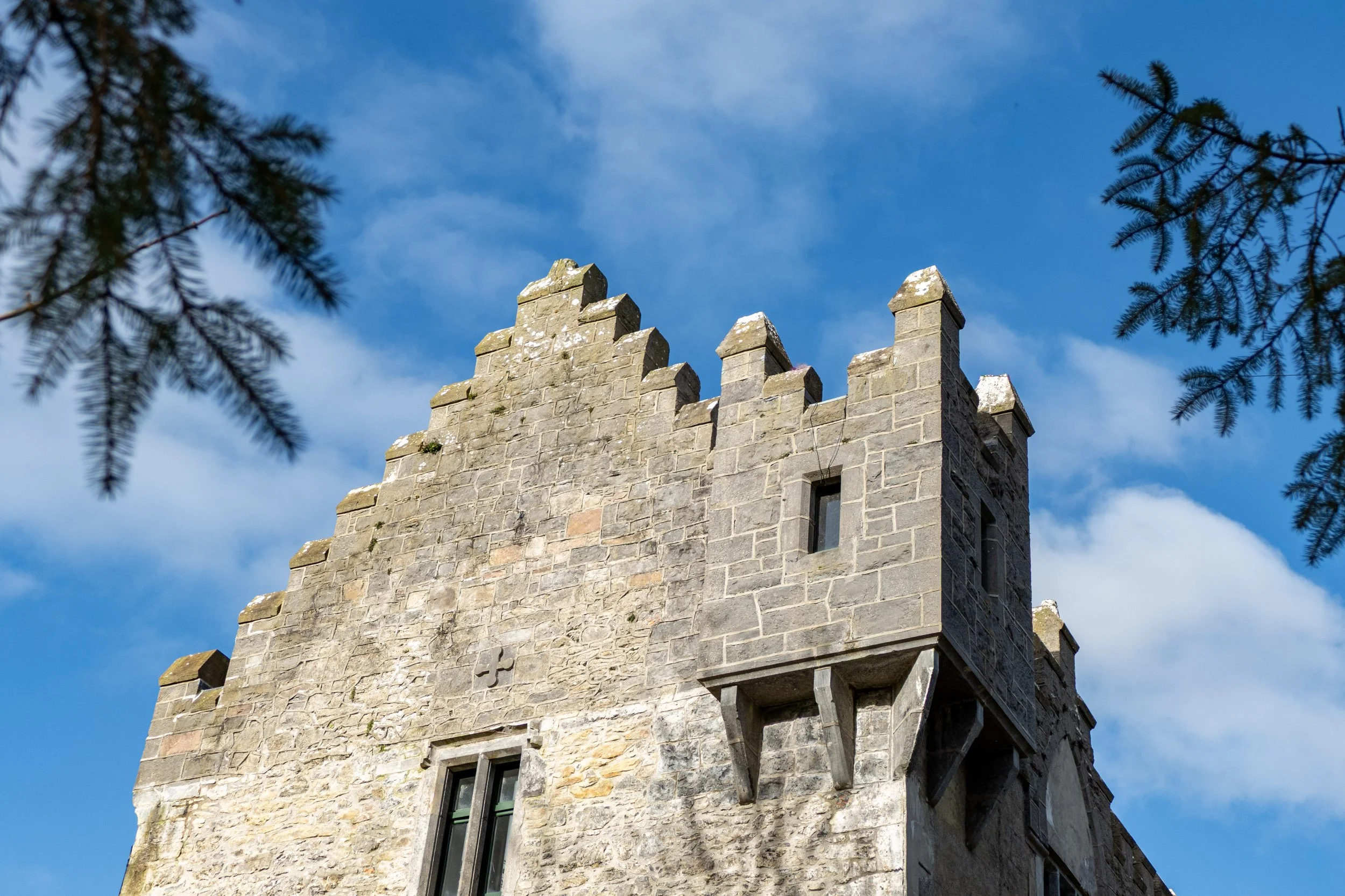 A stone castle tower with battlements, framed by tree branches against a blue sky with some clouds.