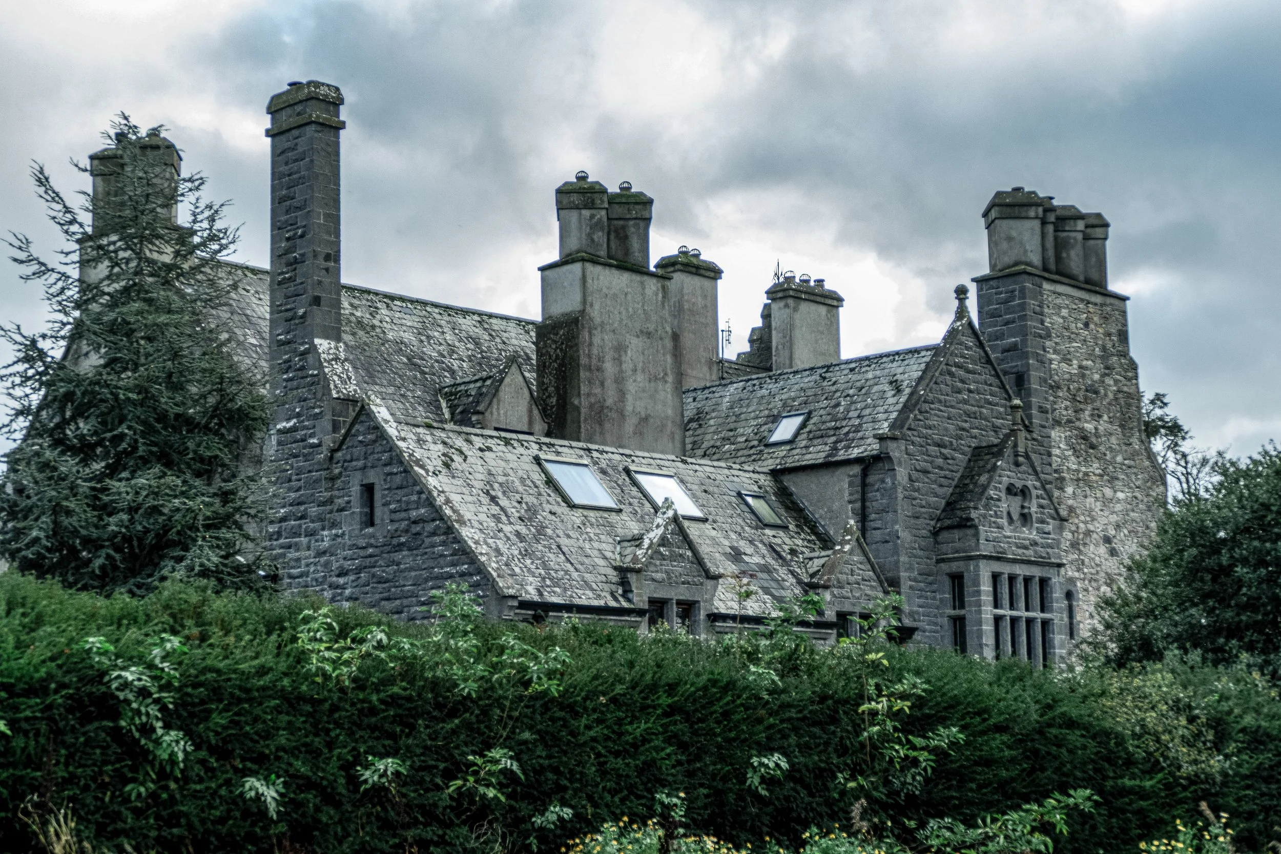 A large, old stone house with multiple chimneys and slate roof, surrounded by green bushes and trees under a cloudy sky.