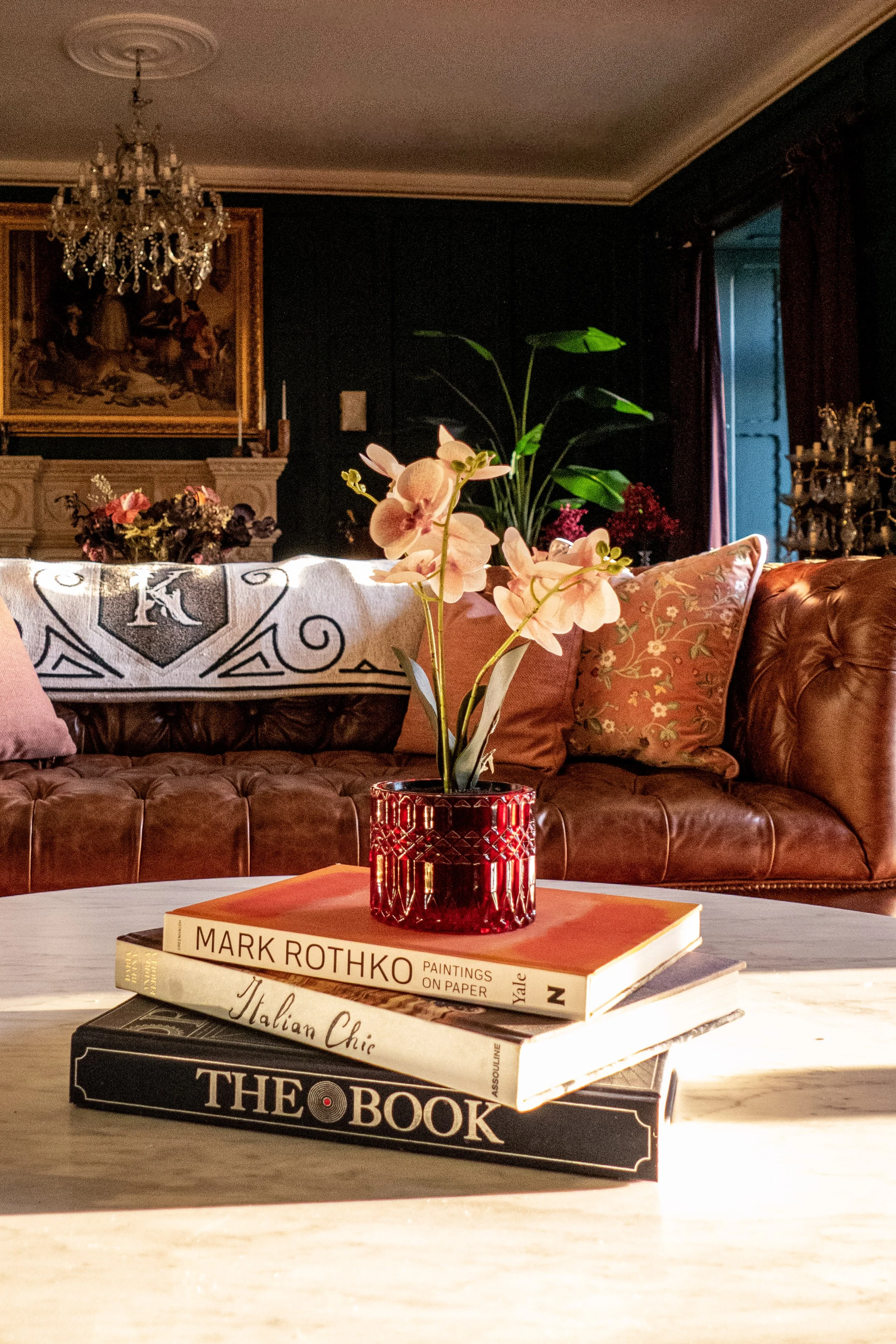 A close-up of a table with four stacked books and a pink orchid in a red vase. The background shows a living room with a brown leather sofa, decorative pillows, a patterned throw, and dark walls with artwork and plants.