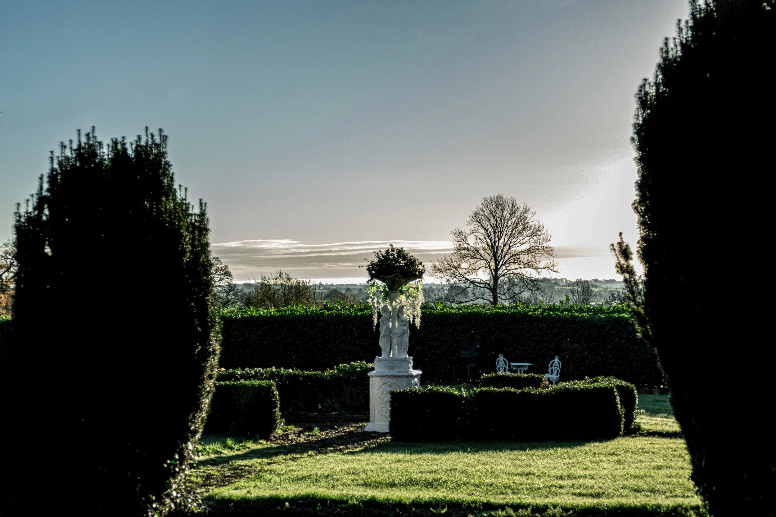 A garden with well-manicured bushes and a white statue of a woman holding a child, framed by two large trees, with a background of trees and a clear sky.