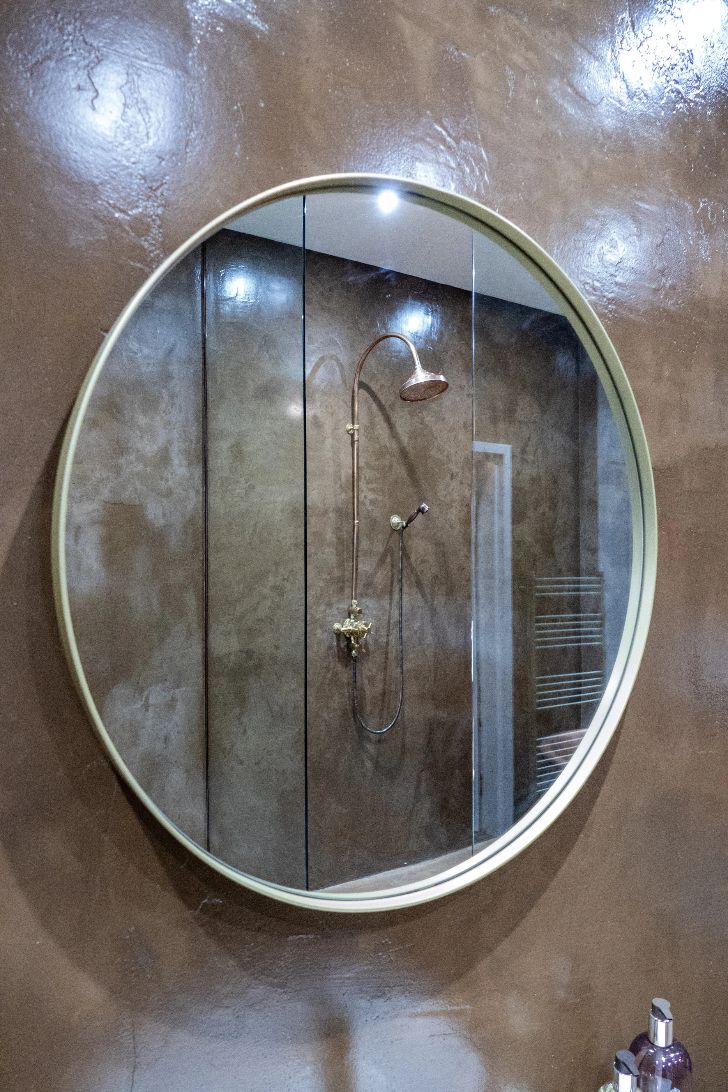 Round mirror reflecting a shower area with a copper showerhead and faucet, built into a brown wall.