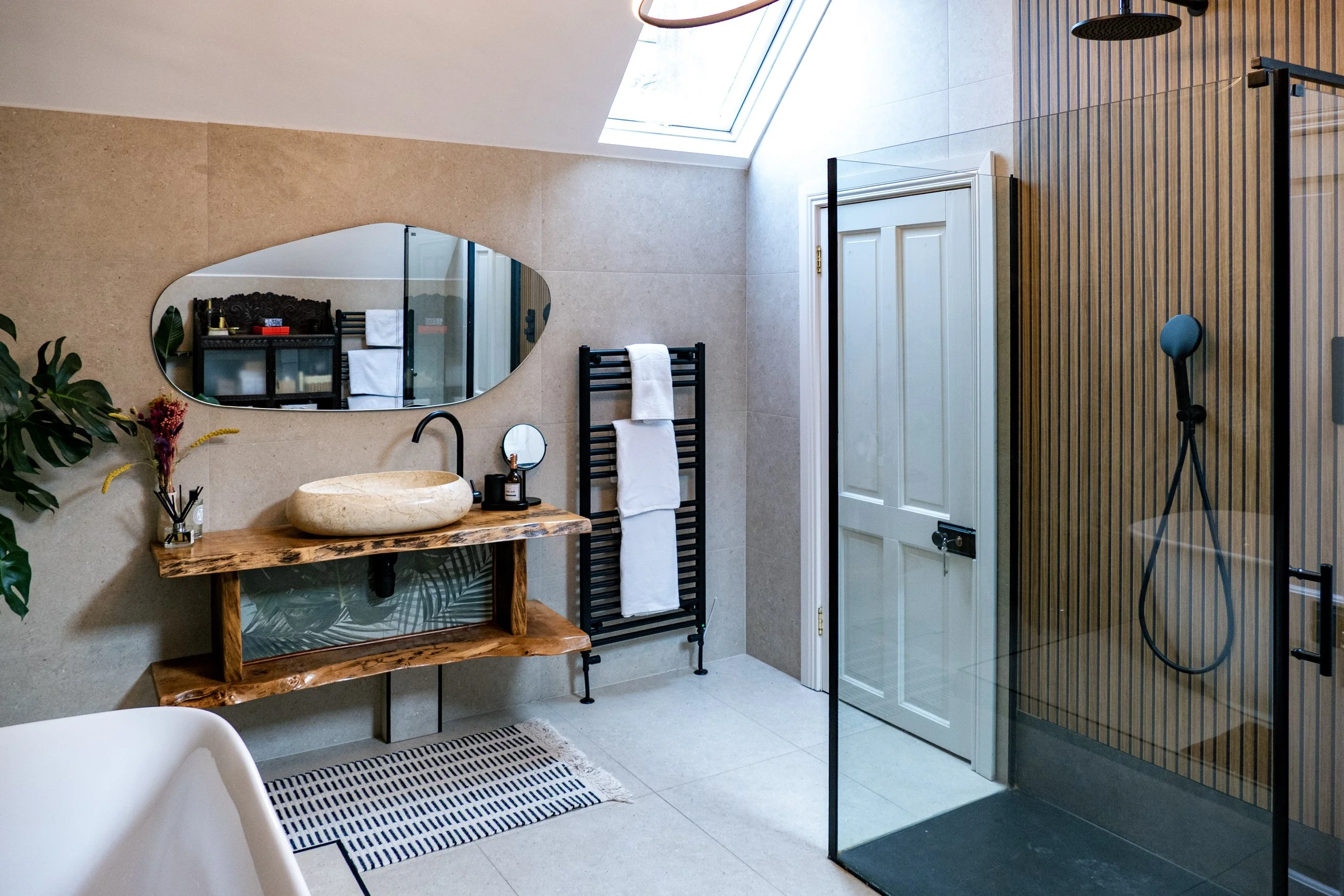 Modern bathroom with a skylight, oval mirror, stone sink on a wooden vanity, black faucet, towel radiator, glass shower enclosure with wooden slat walls, and a white door.