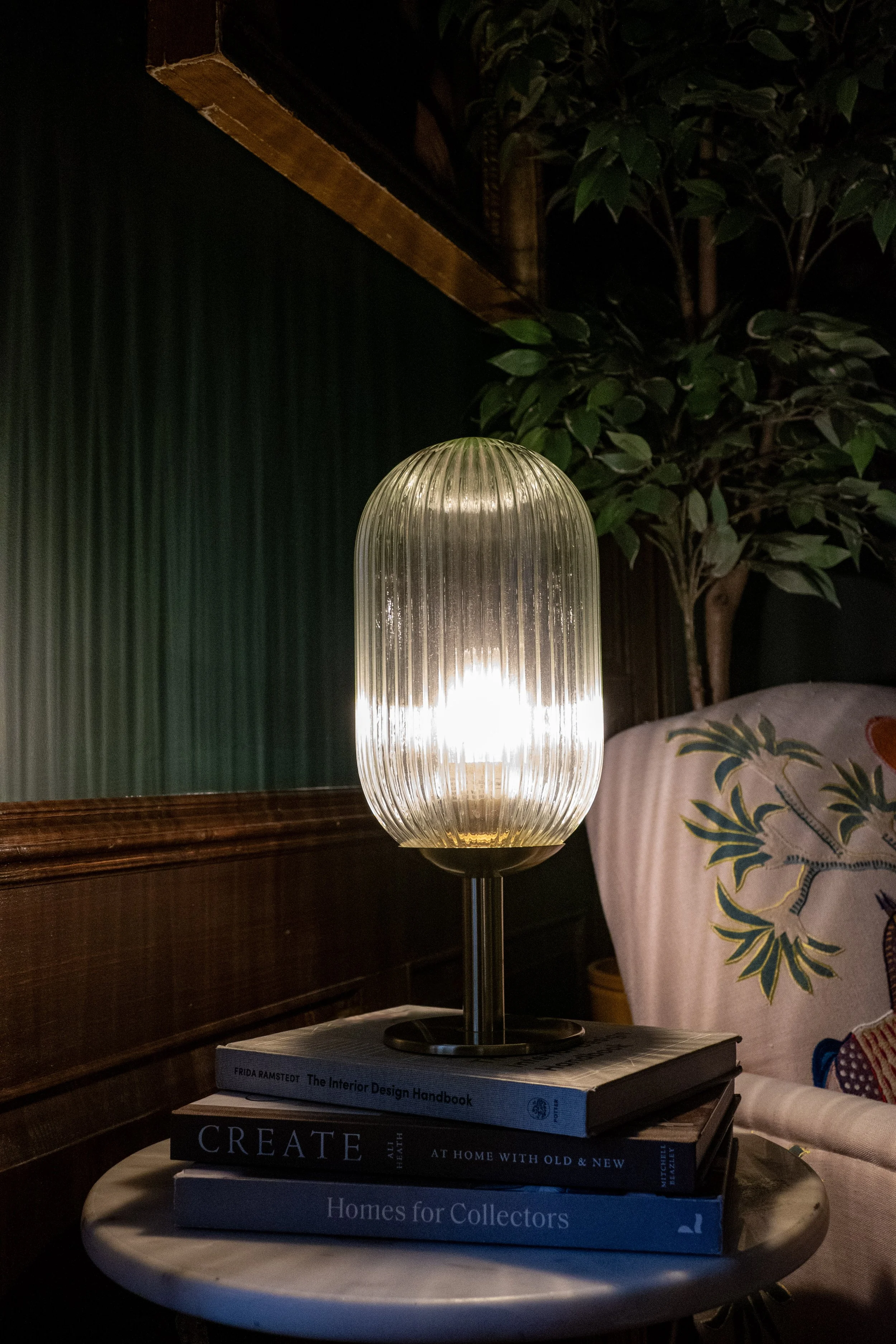 A modern table lamp with a ribbed glass shade on top of a stack of books on a small round table, with a patterned cushion and a green leafy plant in the background.