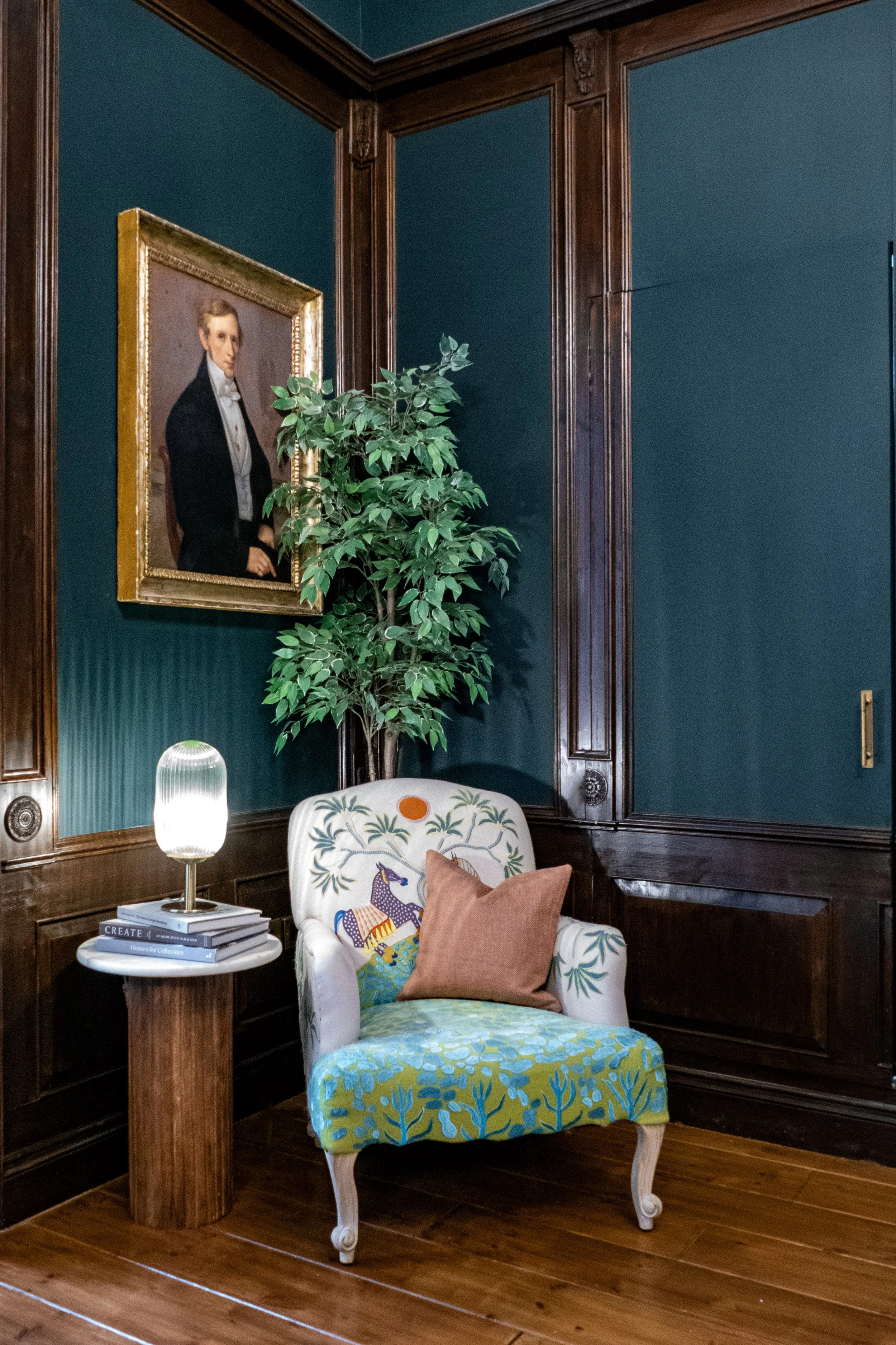 An interior corner with a wooden-framed portrait of a man in formal attire hanging on a dark teal wall, a potted plant, a patterned armchair with a brown pillow, a small side table with a lamp and books, and wooden flooring.