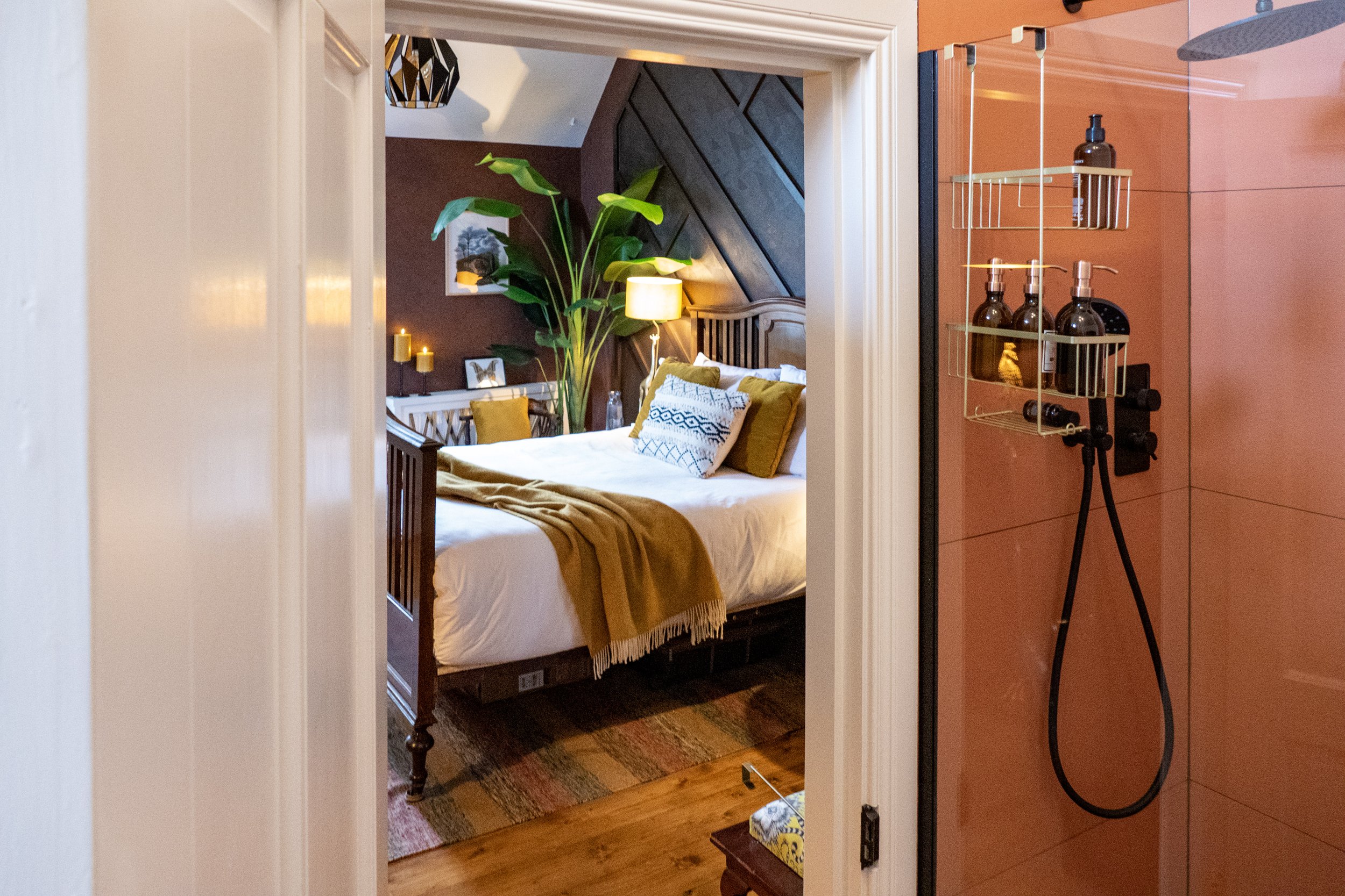 View into a bedroom with a bed, pillows, and a large plant, seen from a bathroom or shower area with pink-tiled walls and a metal shower caddy.