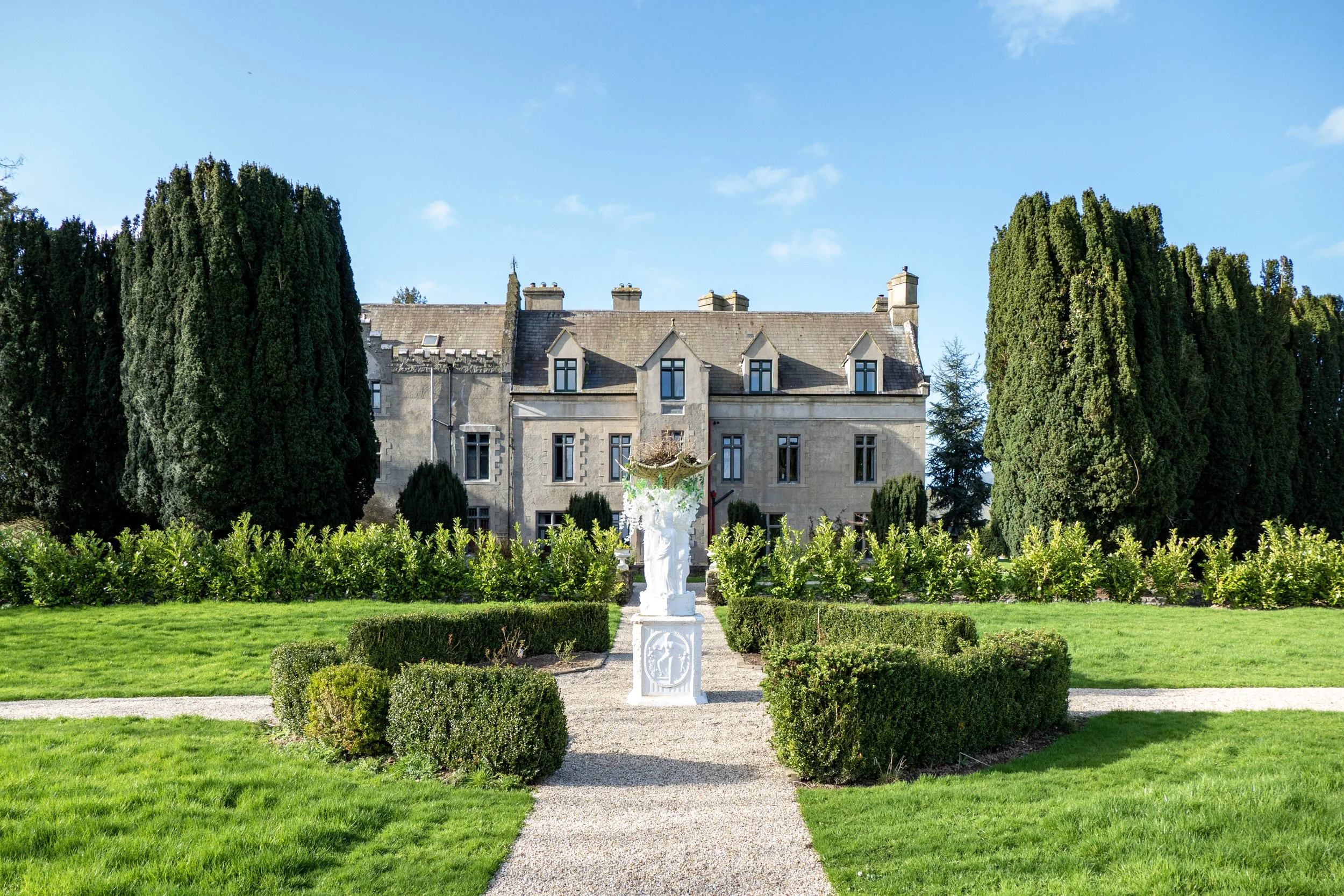 A large historic stone mansion surrounded by a well-manicured garden with symmetrical bushes, tall cypress trees, and a gravel path leading to a white sculpture or statue in the center of the garden, under a blue sky.