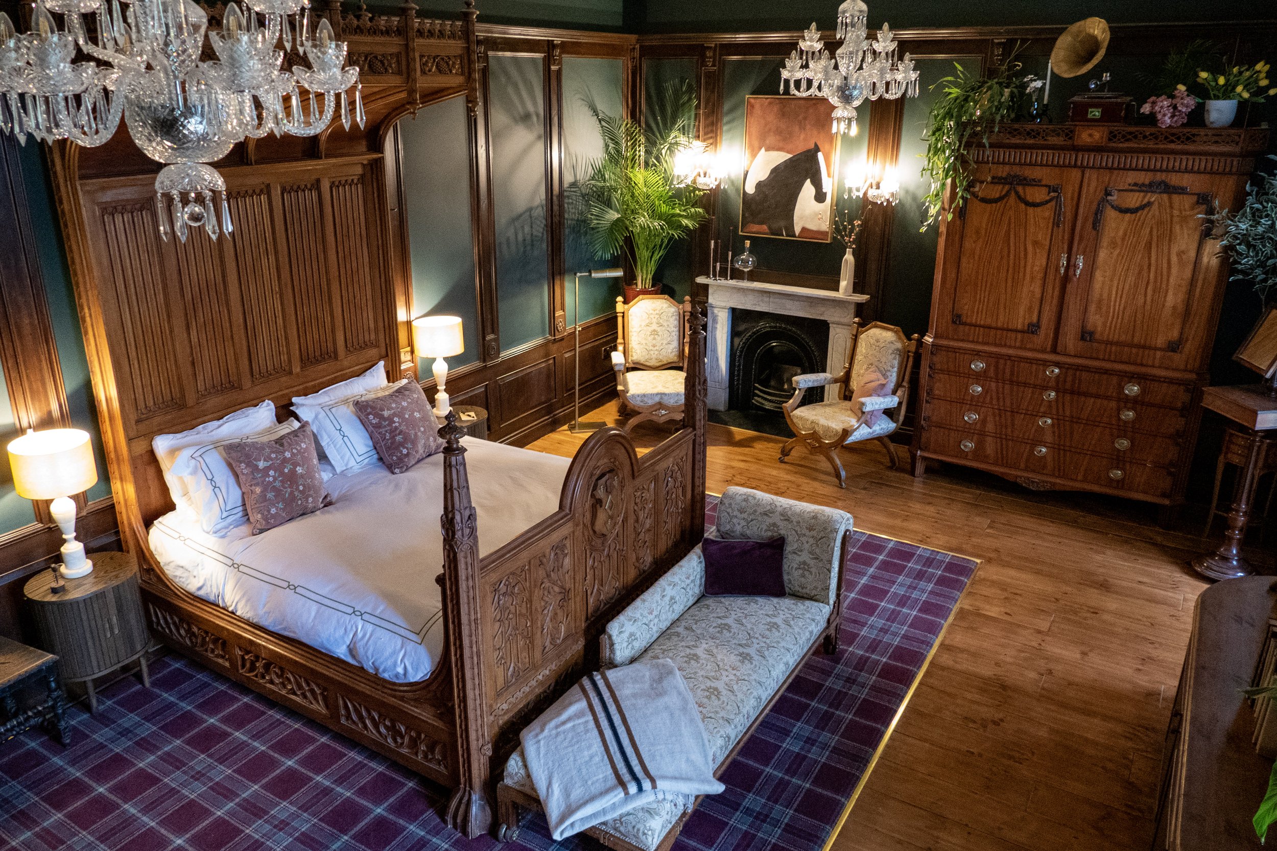 A luxurious bedroom with dark green walls, ornate wooden paneling and furniture. There is a four-poster bed with white bedding and decorative pillows, two side tables with lamps, a fireplace with two upholstered chairs, a large wooden wardrobe, and a
