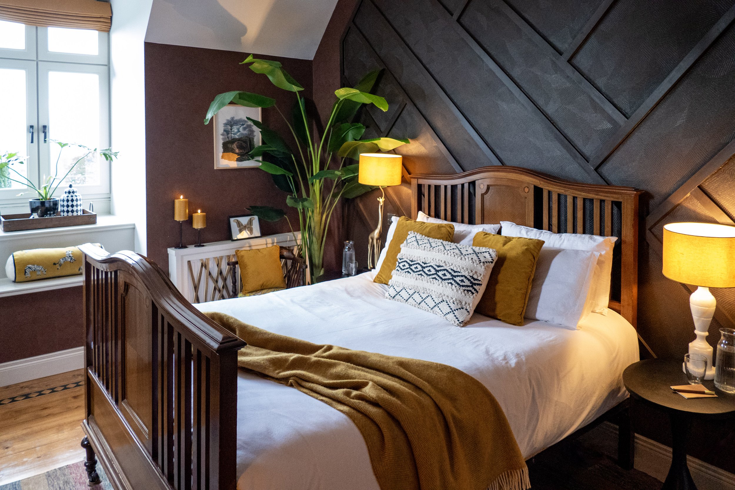 A bedroom with a wooden bed, decorative pillows, yellow throw blanket, large green plants, bedside lamps, a window with potted plants, and wall art.