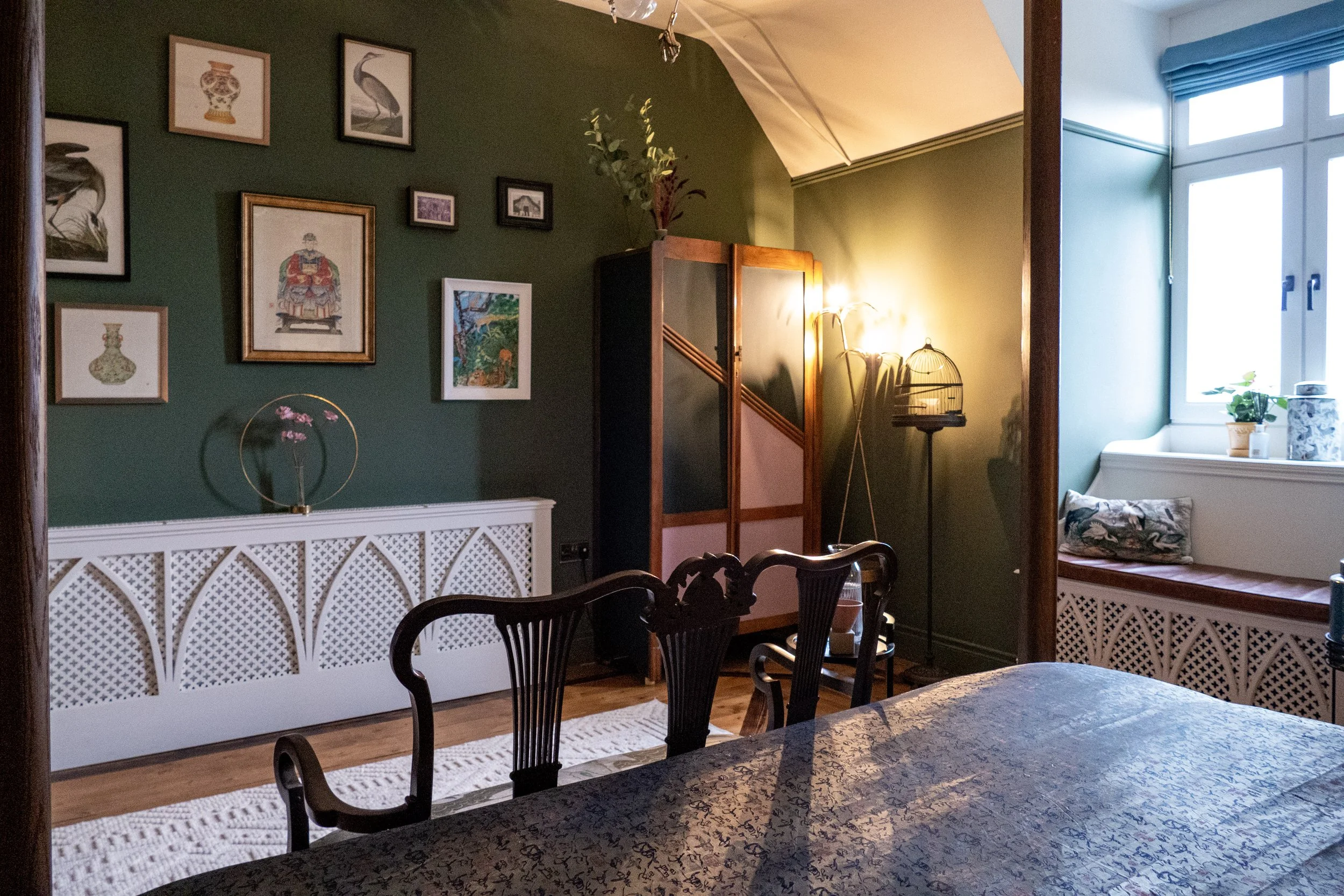 Living room with green and yellow walls, decorated with framed art and plants, a wooden room divider, corner window seat with cushions, and a dining table with chairs in the foreground.