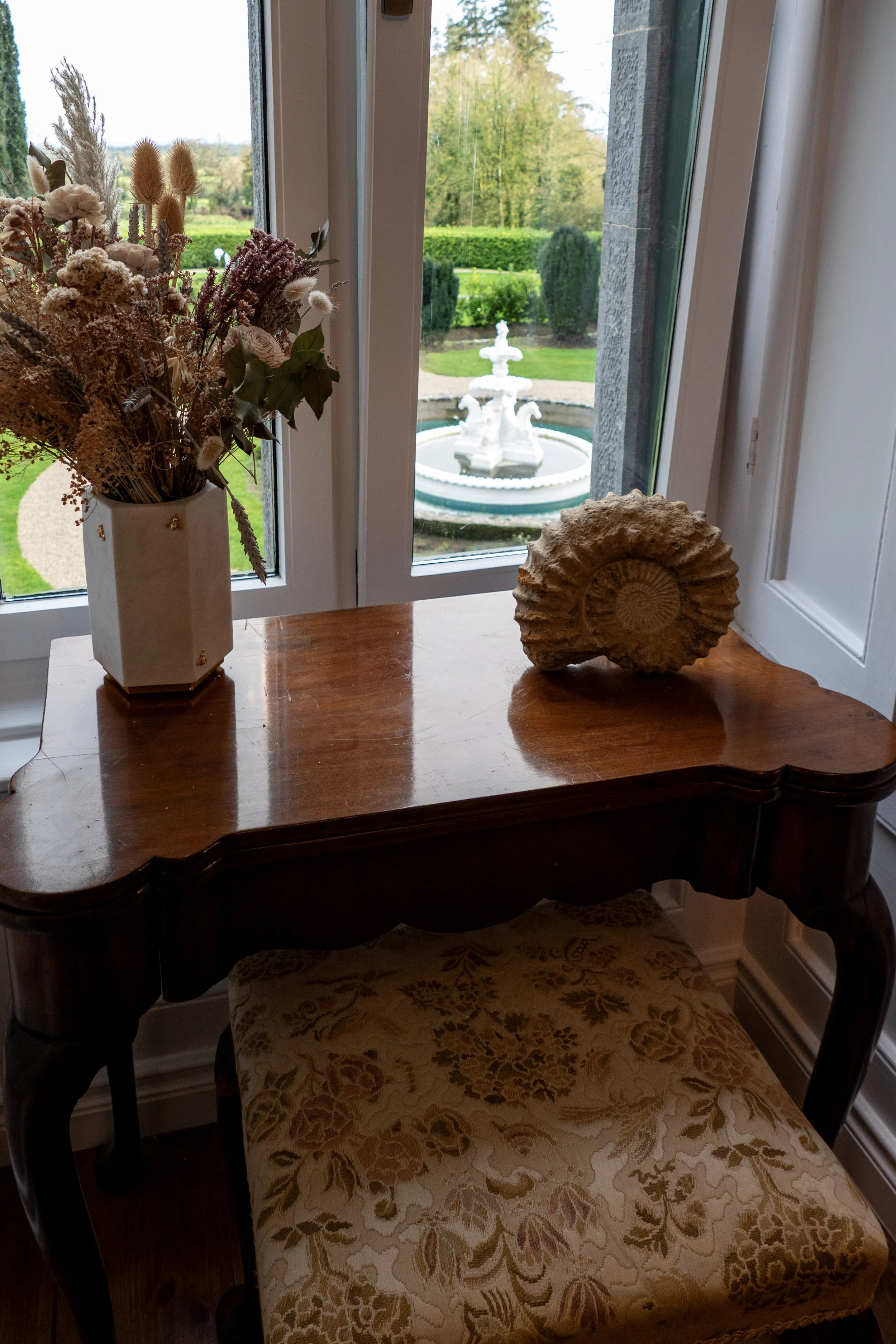 A wooden table by a window with a vase of dried flowers and a large seashell on it. Outside the window, a white fountain is in a garden with green trees and shrubs.