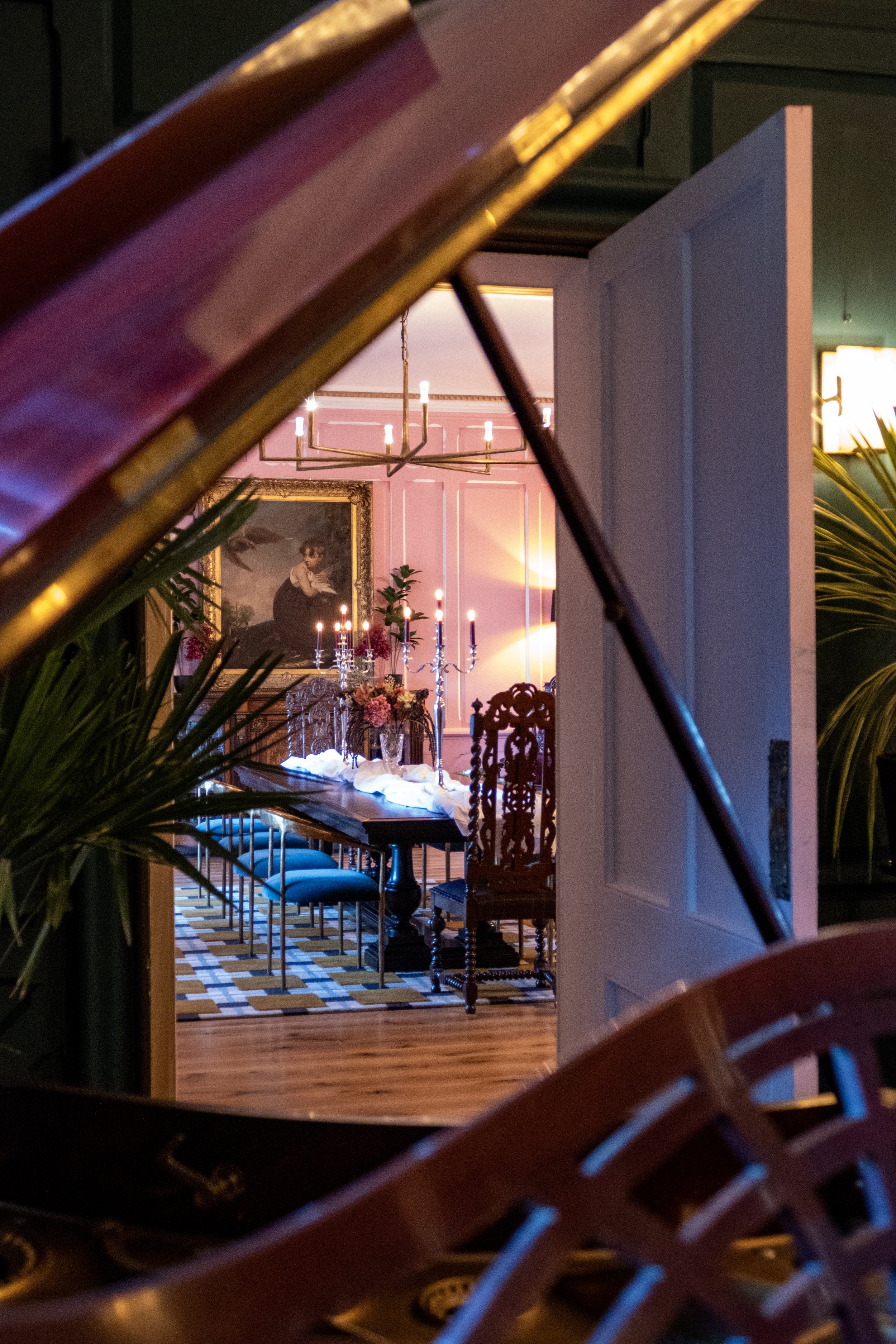 View through the curved corner of a piano showing an elegant dining room with a long table, blue chairs, pink walls, a chandelier, candelabras, floral centerpieces, and a large portrait.