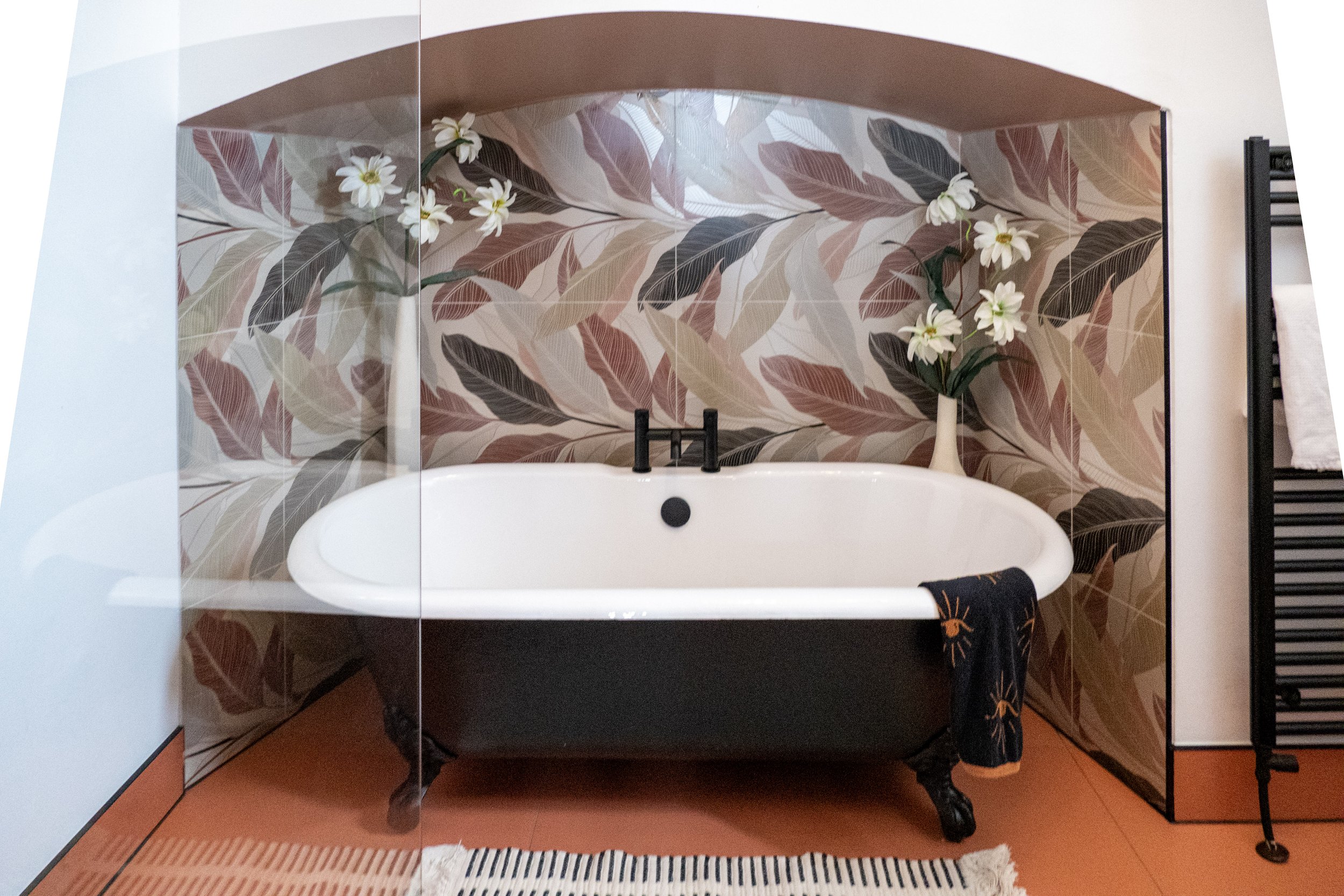 A vintage clawfoot bathtub with a black exterior and white interior, decorated with a dark towel hanging over the side, in a bathroom with a tropical leaf-patterned wallpaper, a vase with white flowers, a black towel rack, and a striped rug.
