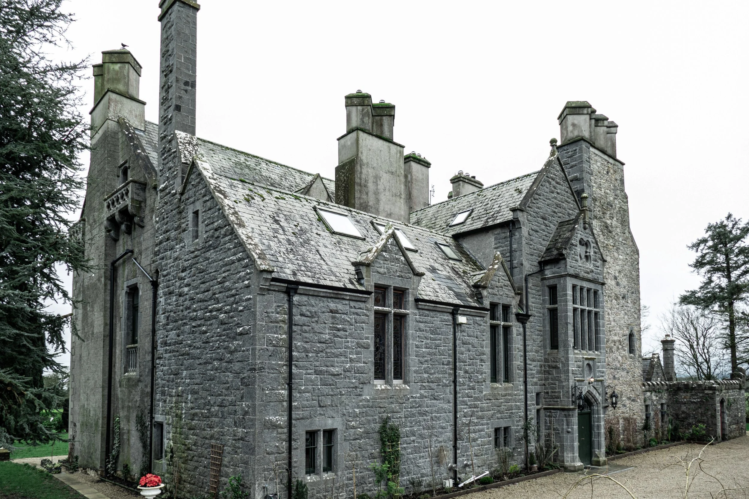 A historic stone castle with multiple chimneys, gothic windows, and a sloped roof, surrounded by trees and a gravel pathway.