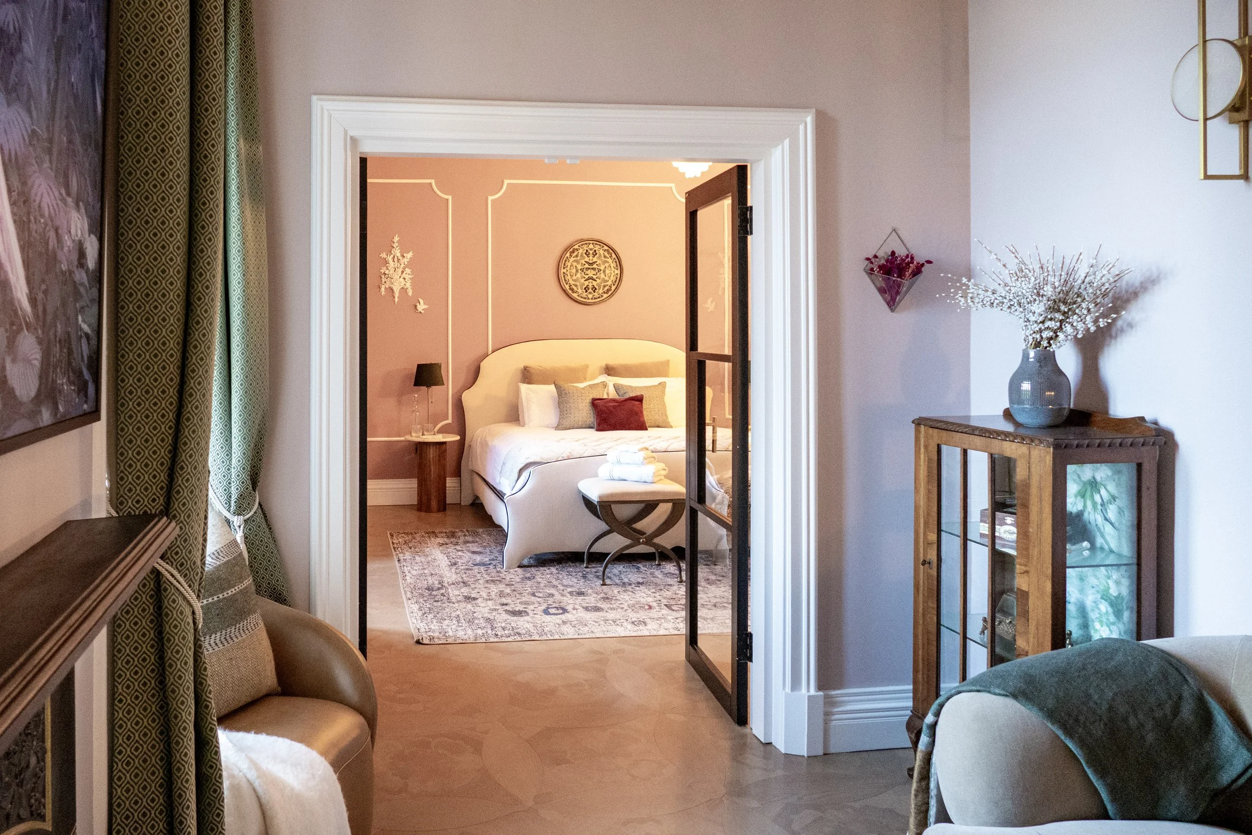 Interior view of a hotel room with a bedroom visible through a doorway. The bedroom has a bed with white linens and multiple pillows, a nightstand with a lamp, and decorative wall art. The foreground shows part of a sitting area with a beige armchair