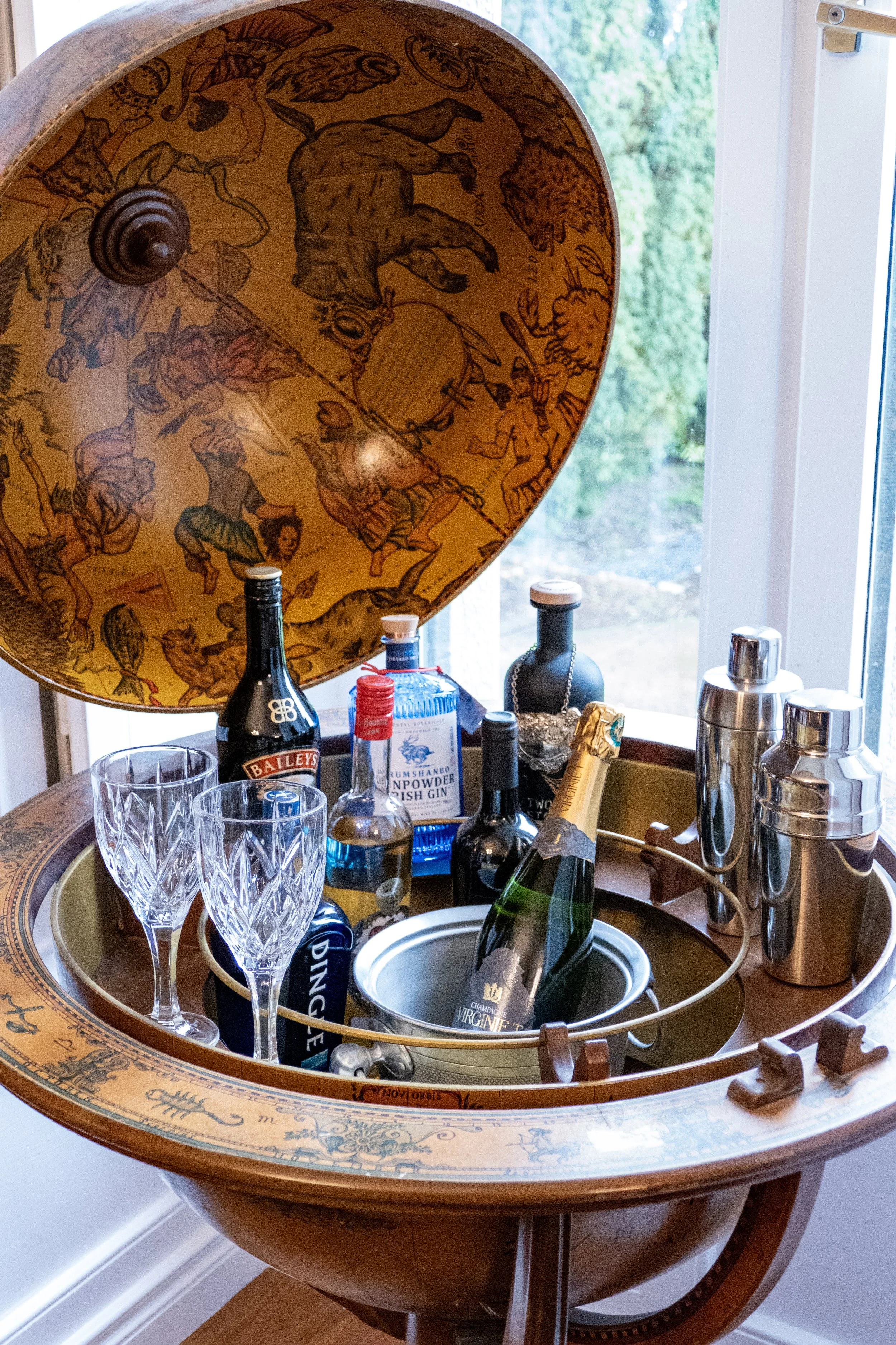 A vintage globe bar cart with a celestial map design on the interior lid, opened to reveal bottles of alcohol, glasses, and bar accessories inside.