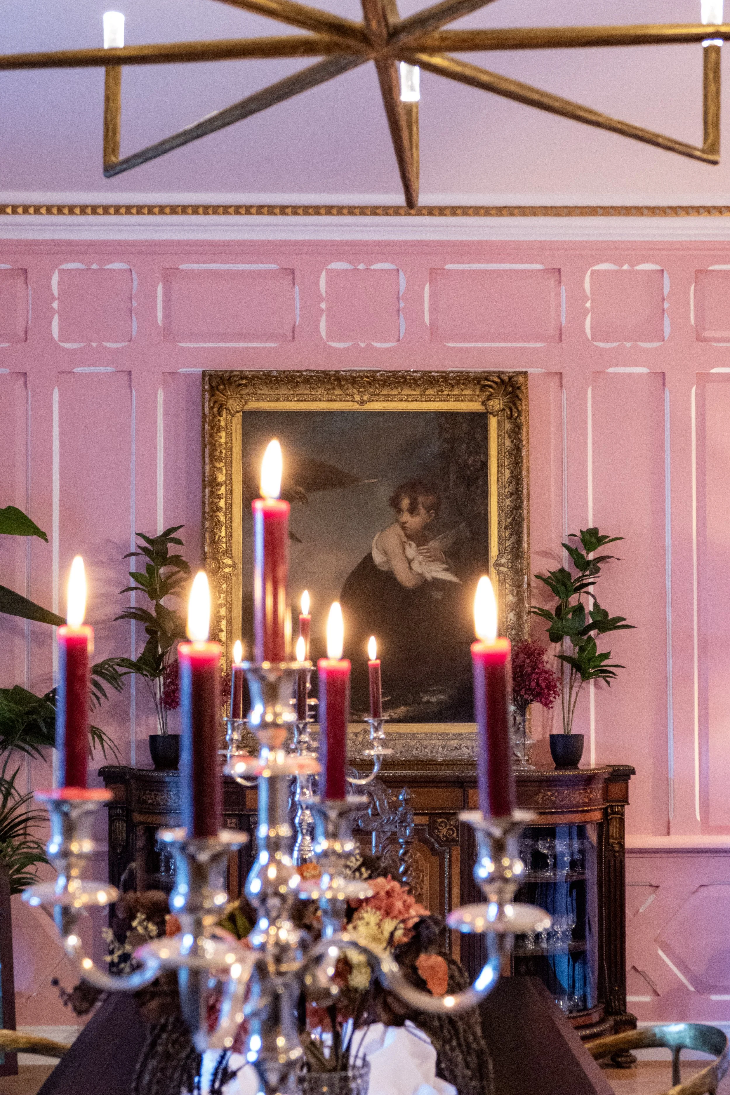 A chandelier with lit red candles hangs above a dining table with a candelabrum, set against a pink paneled wall with a framed painting of a young girl holding a feather.