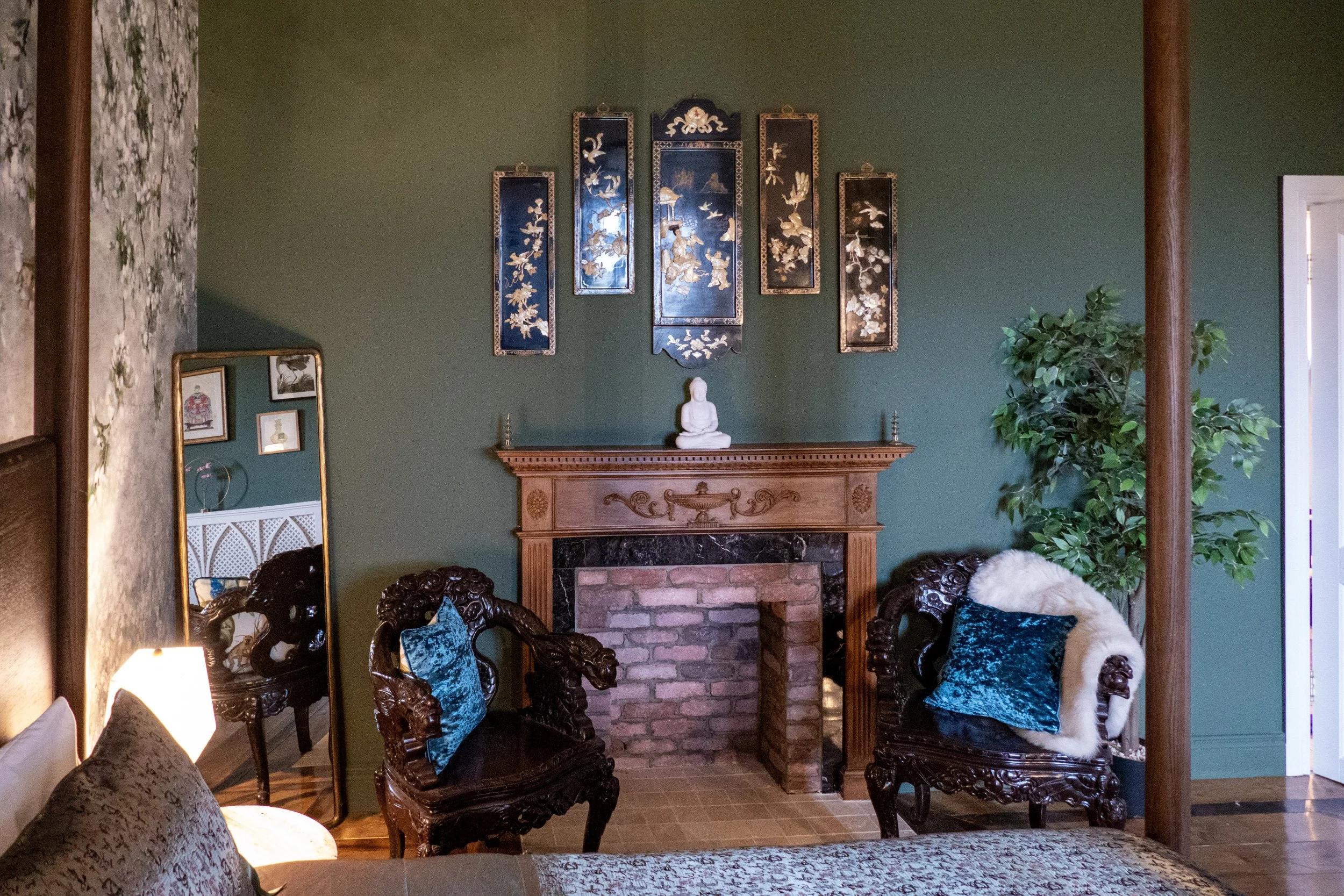 Living room with green walls, ornate wooden fireplace mantel, decorative wall art, a white Buddha statue, two wooden armchairs with blue pillows, a large leafy plant, a mirror, and a portion of a bed with a patterned pillow.