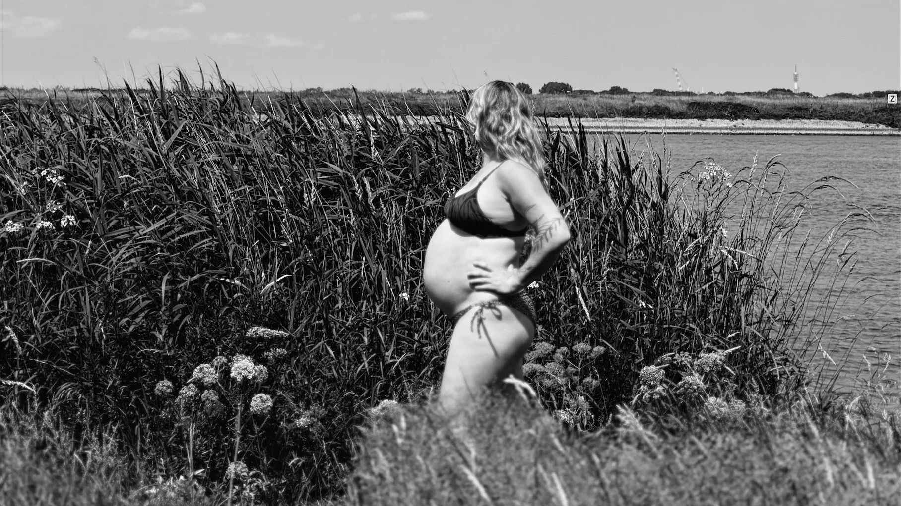 Zwangere vrouw met tatoeages in bikini staat aan de rand van een waterlichaam, omgeven door hoge planten en gras, tegen de achtergrond een open landschap met blauwe lucht.