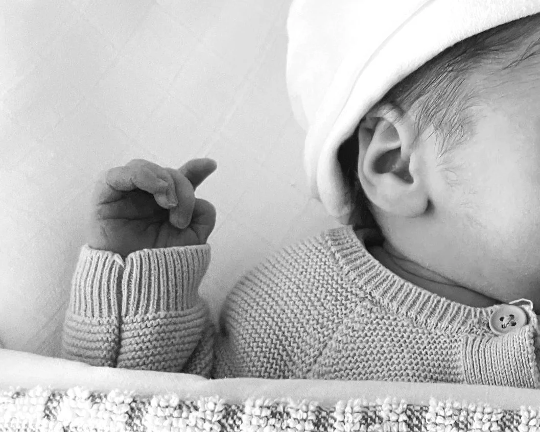 Close-up van een baby met mutsje, gekleed in een geribde trui, met een hand omhoog.
