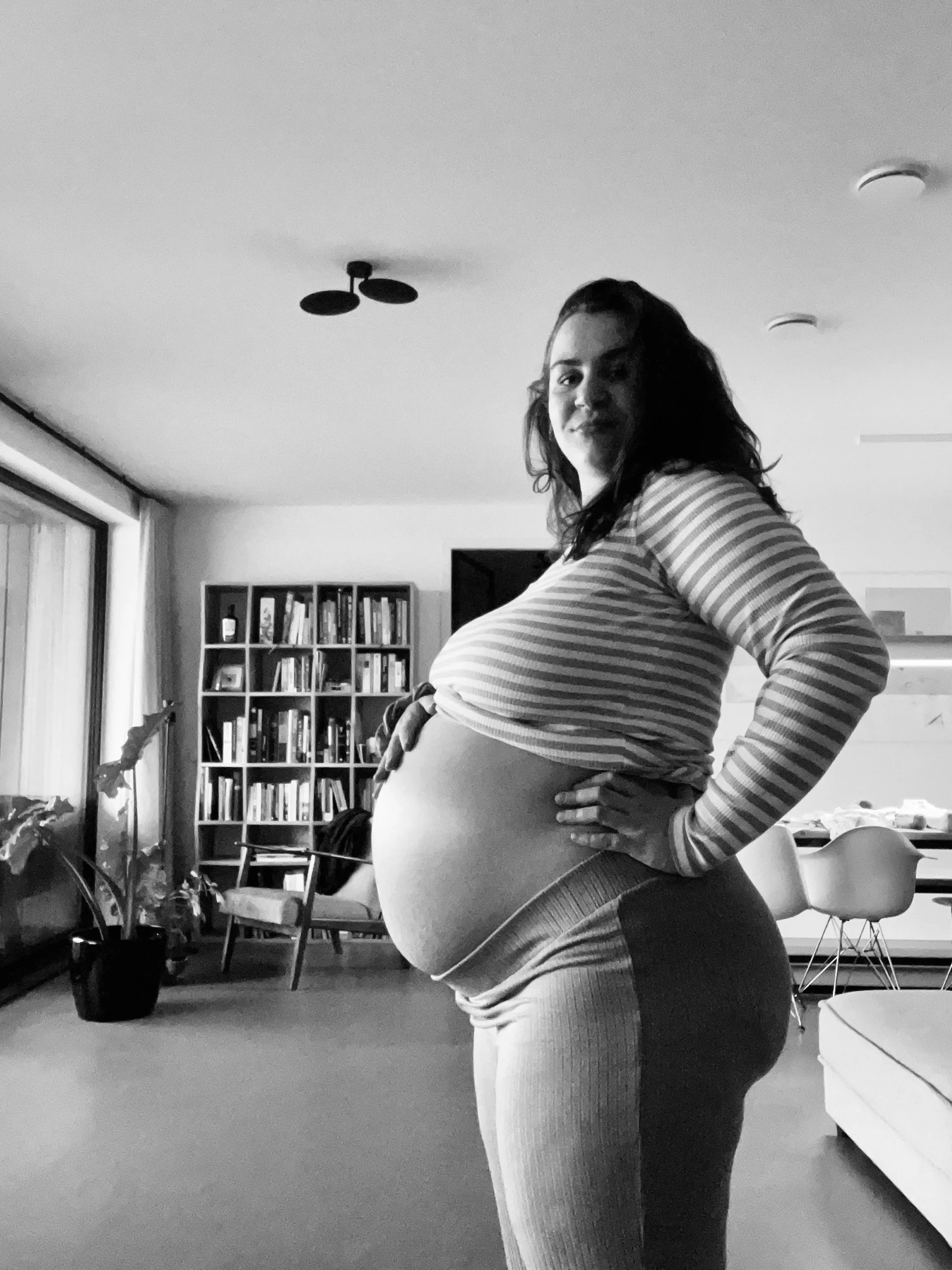 Zwangere vrouw met een brede buik in een woonkamer, zittend op een stoel, met boekenplank op de achtergrond, in zwart-wijn