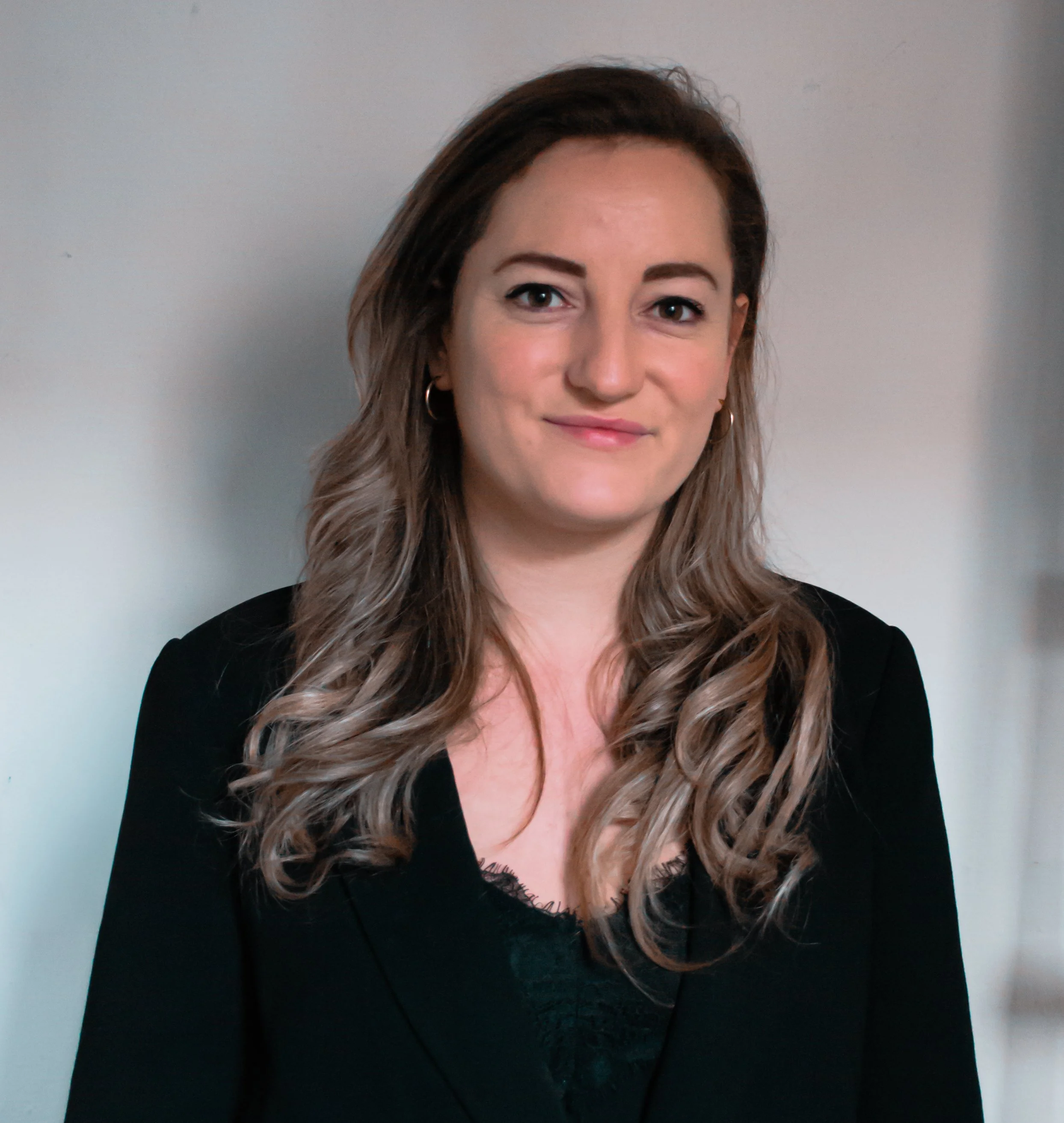 Portret van een jonge vrouw met lang, golvend, bruin haar en lichte make-up, gekleed in een zwarte top, staand voor een lichtgrijze achtergrond.