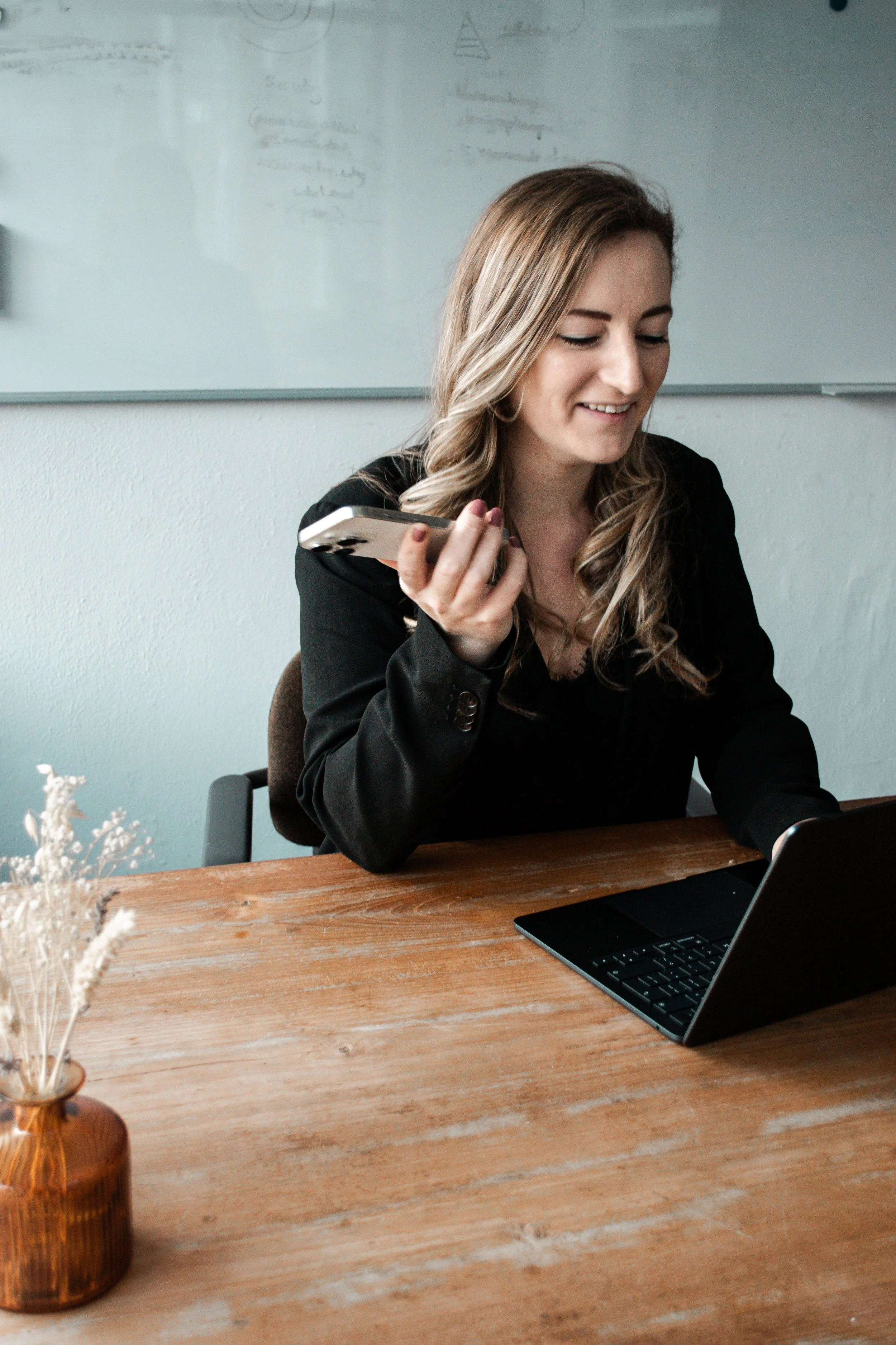 Een vrouw met lang, lichtbruin haar die glimlacht terwijl ze op haar smartphone kijkt, zittend aan een houten tafel met een open laptop voor haar, in een kantooromgeving met een whiteboard op de achtergrond.