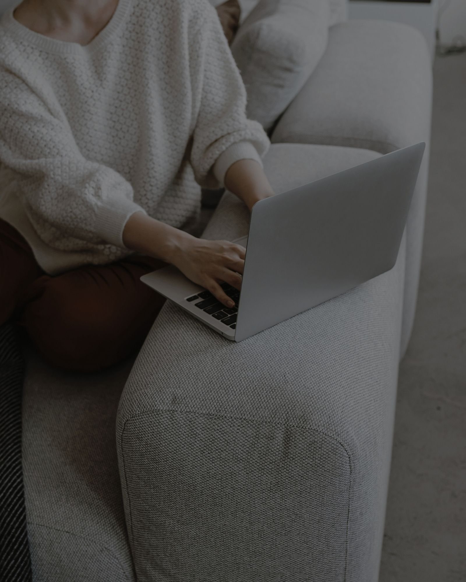 Persoon die op een bank zit met een open laptop op schoot, gekleed in een beige gebreide trui, in een woonkamer.