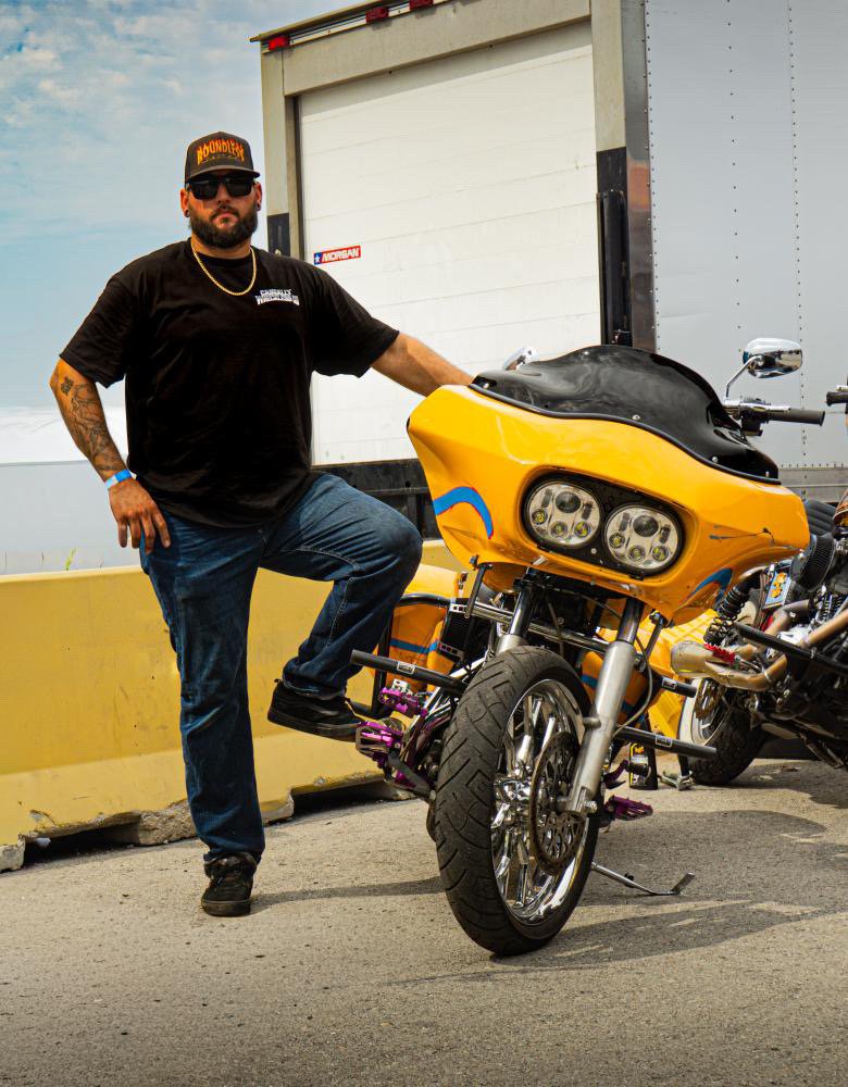 A man with a beard wearing sunglasses, a black t-shirt, jeans, and a cap standing next to a custom yellow motorcycle with black accents and blue details.