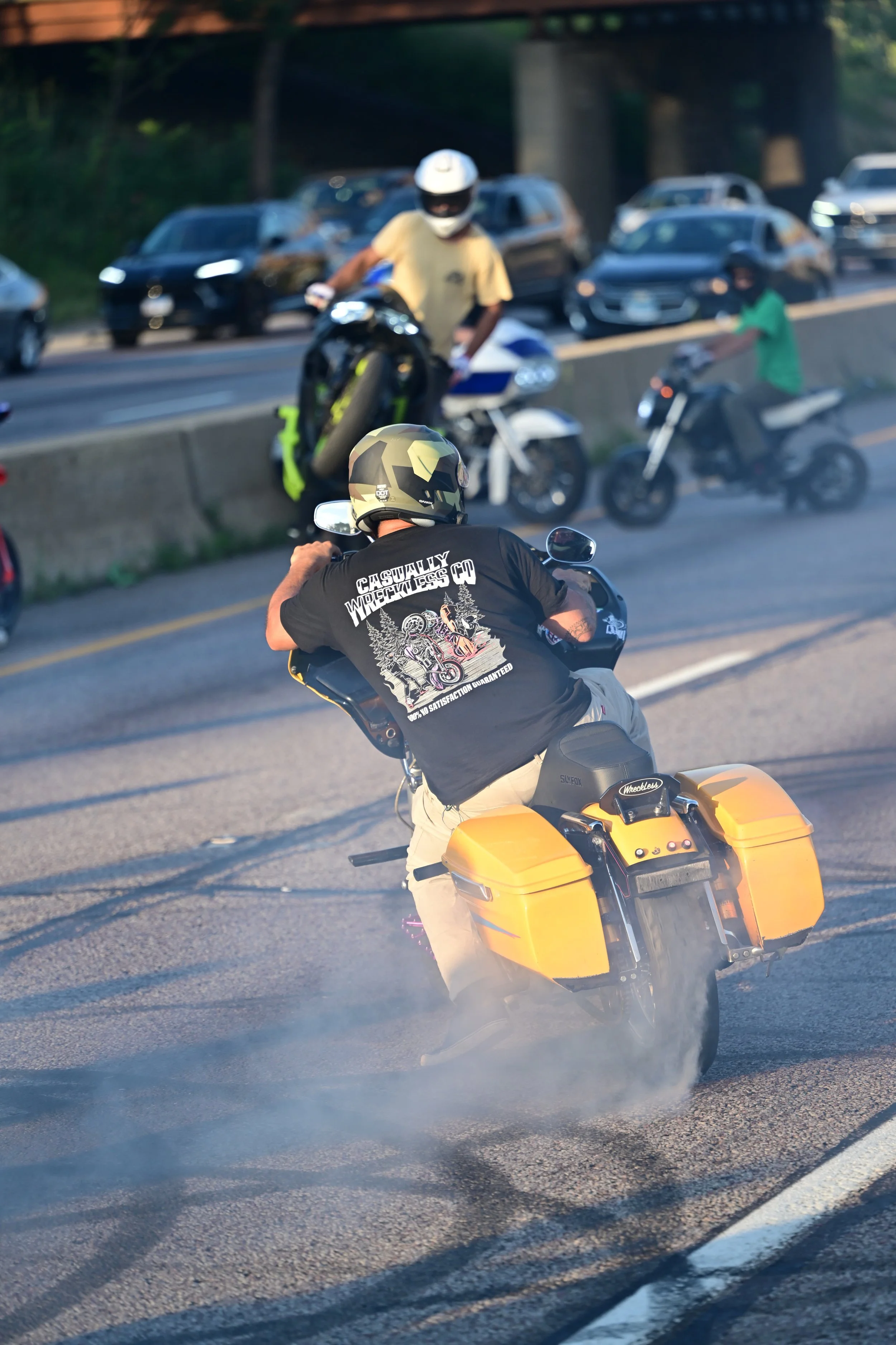 Motorcycle riding on a highway with smoke coming from rear tire, with multiple cars and bikers in background.