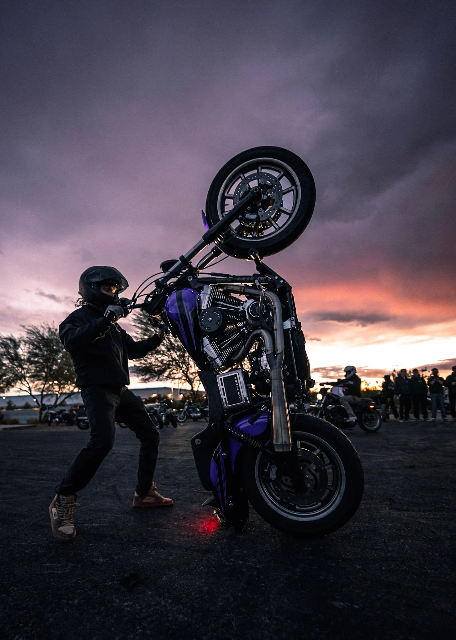 Motorcycle stunt rider performing a wheelie at sunset, with a crowd of onlookers and motorcycles in the background