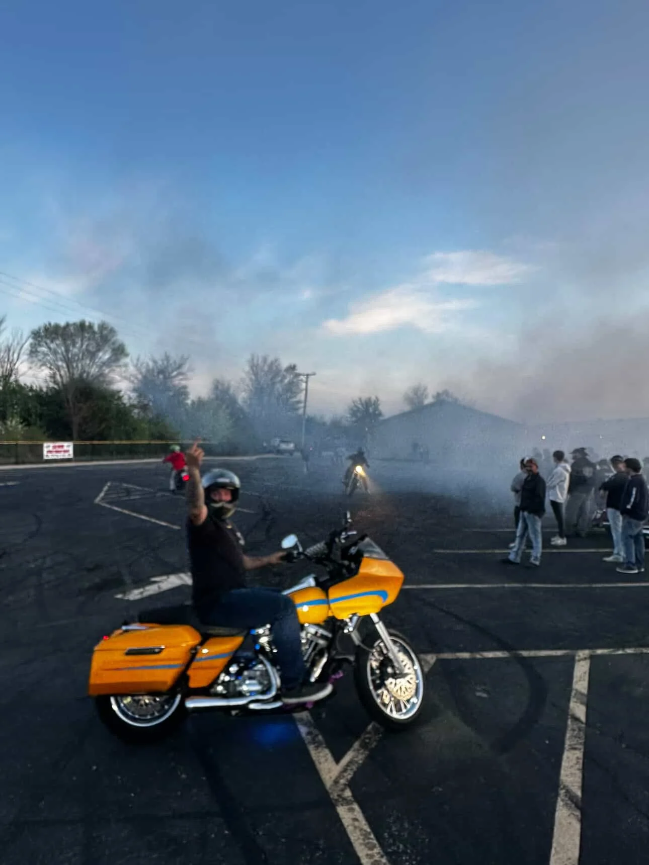 A person wearing a helmet and face mask sitting on an orange and black motorcycle in a parking lot. In the background, there are people standing and observing, some motorcycles, smoke, and a partly cloudy sky.