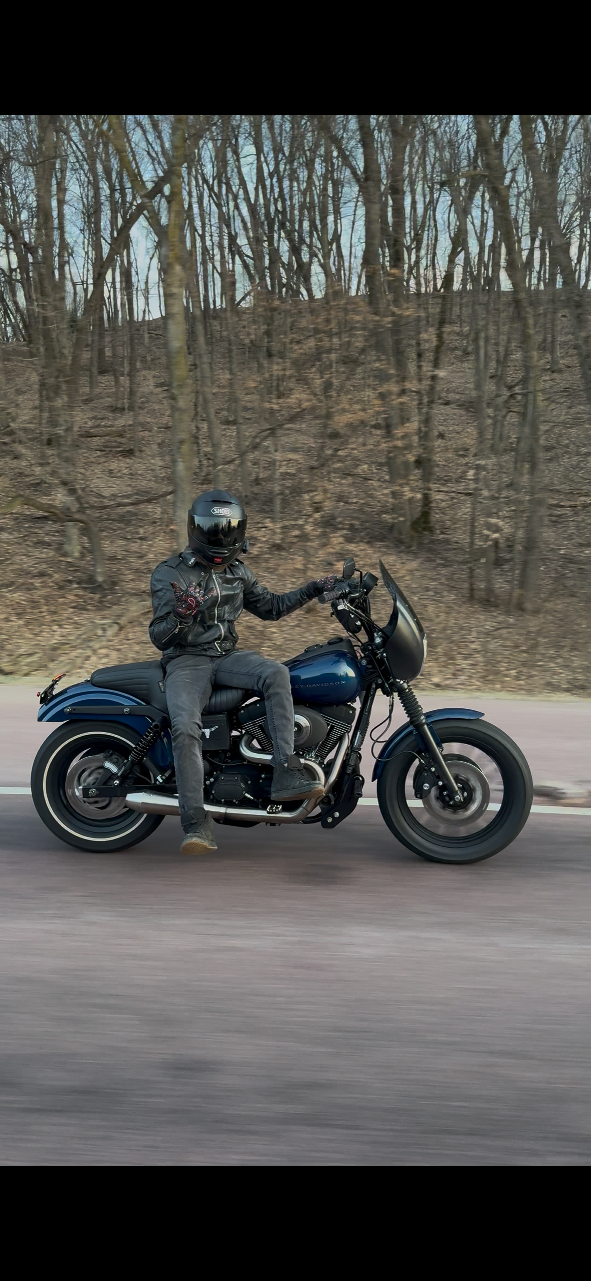Motorcyclist wearing a black helmet and leather jacket riding a black Harley Davidson motorcycle on a road with a wooded background.