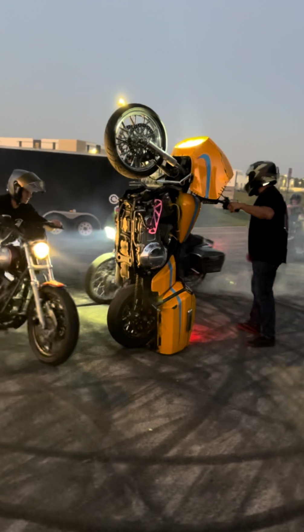 Two motorcyclists standing next to a flipped yellow and red robot-like structure resembling a robot partially upside down, with smoke and light effects in the background during dusk.