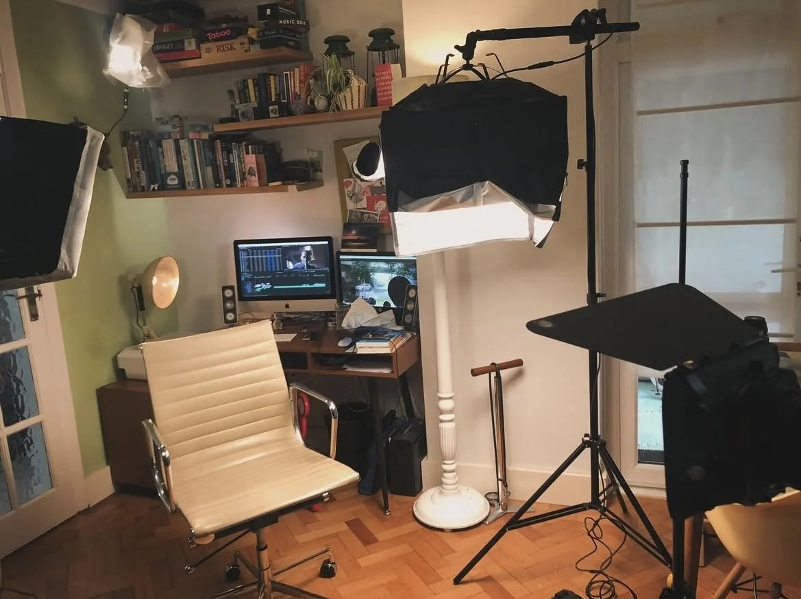 A home office or studio setup with lighting equipment and a computer desk, featuring multiple bookshelves, a white swivel chair, and recording or video editing equipment.