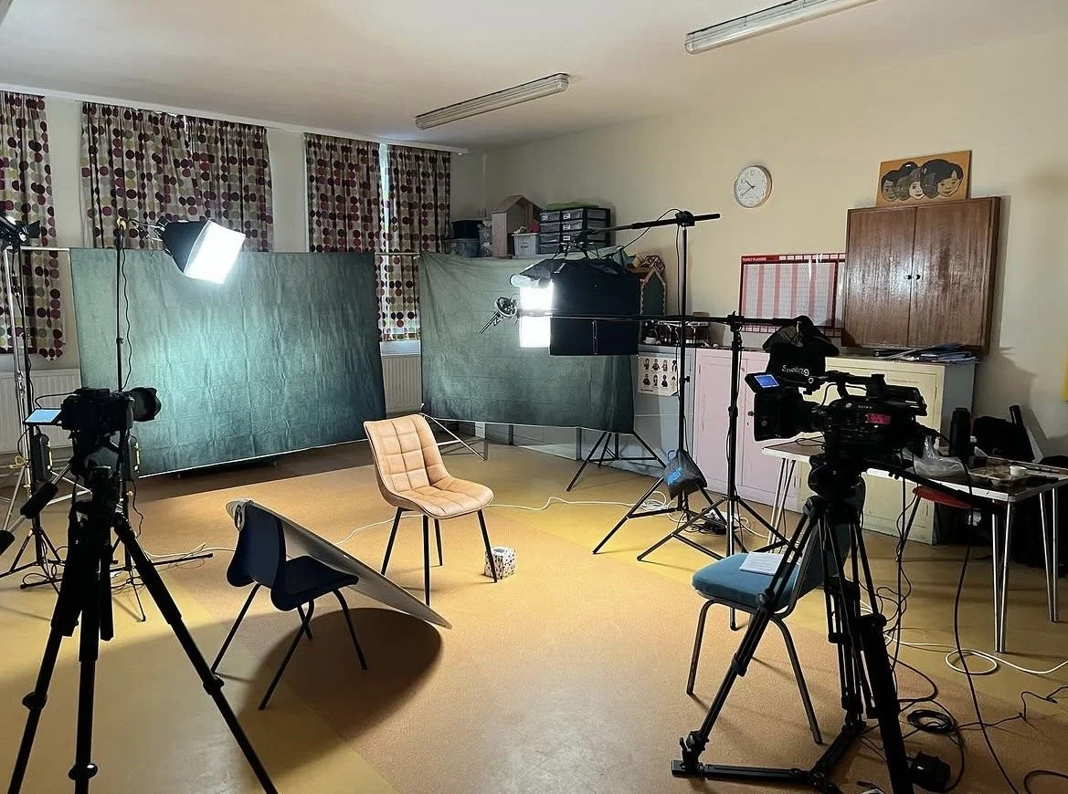 Video production studio with cameras, lighting, chairs, and green screen setup in a room with patterned curtains and a closed wooden cabinet.