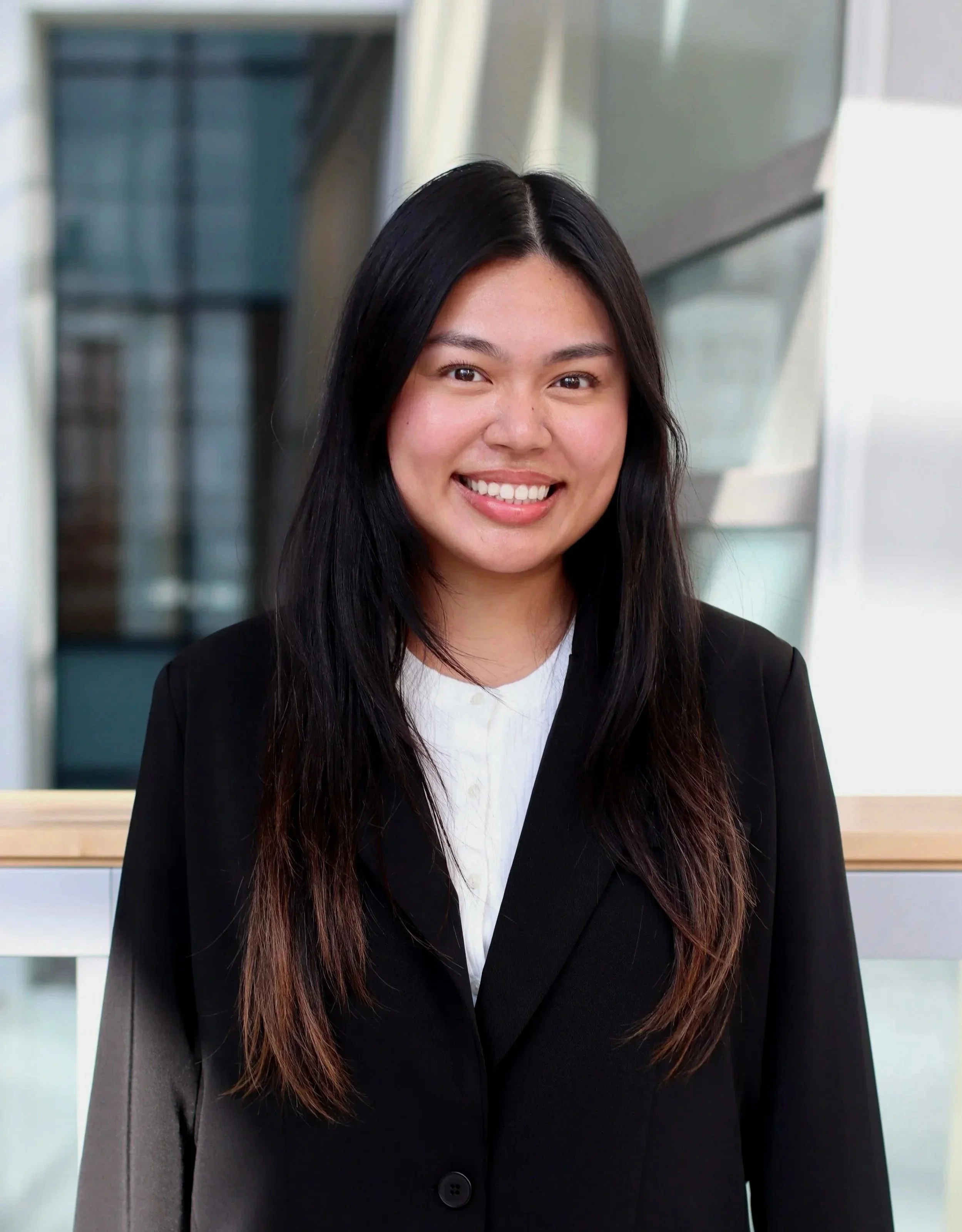 A young woman with long dark hair, wearing a black blazer and white shirt, standing indoors with a background of modern glass architecture, smiling at the camera.
