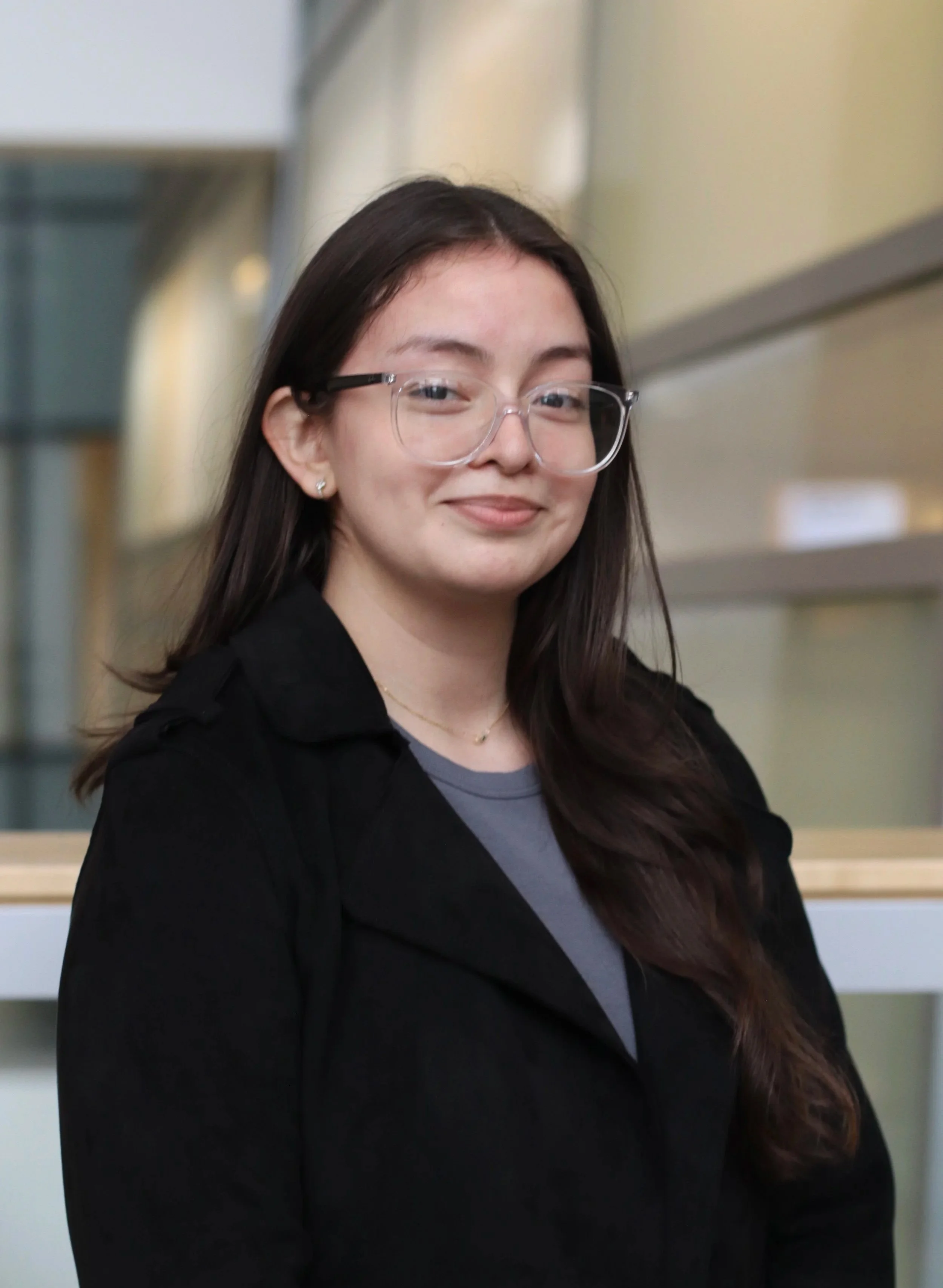 Portrait of a young woman with long dark hair, glasses, earrings, a gray shirt, and a black jacket, smiling in an indoor setting.
