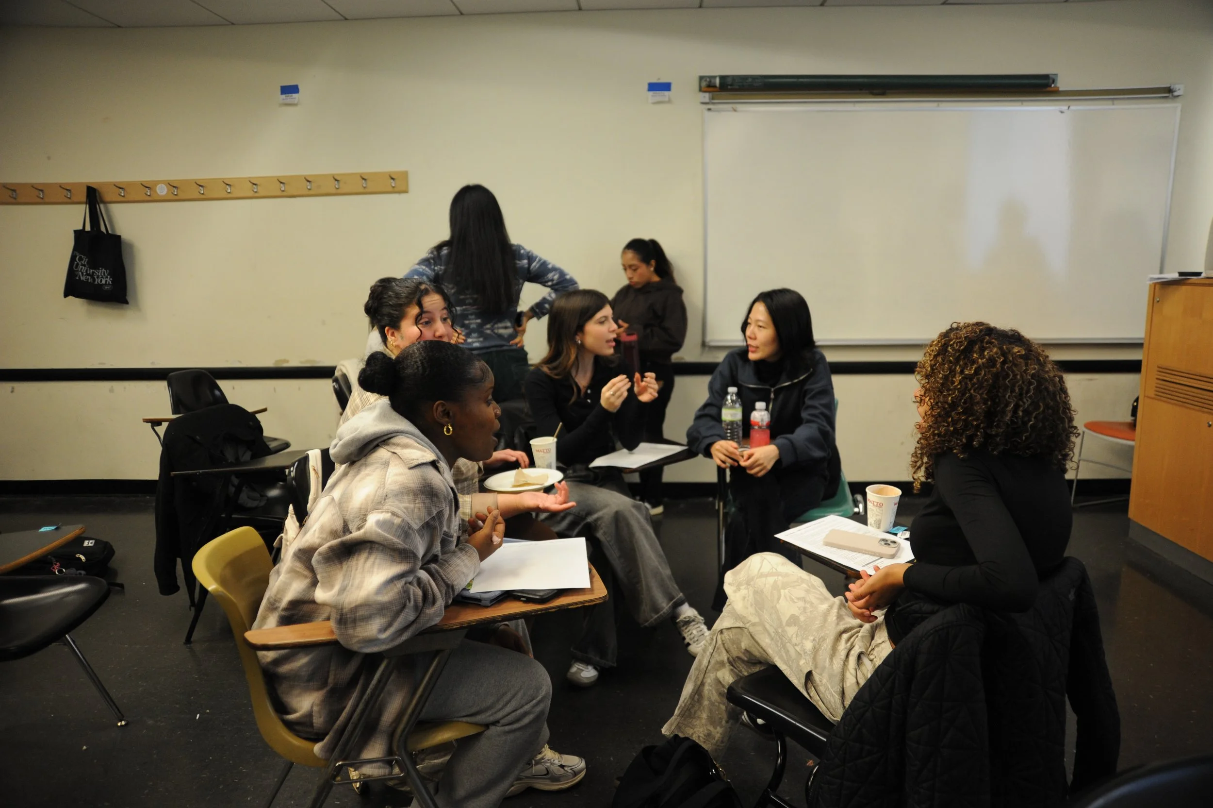 Diverse group of students discussing in a classroom with a whiteboard, desks, papers, and drinks.