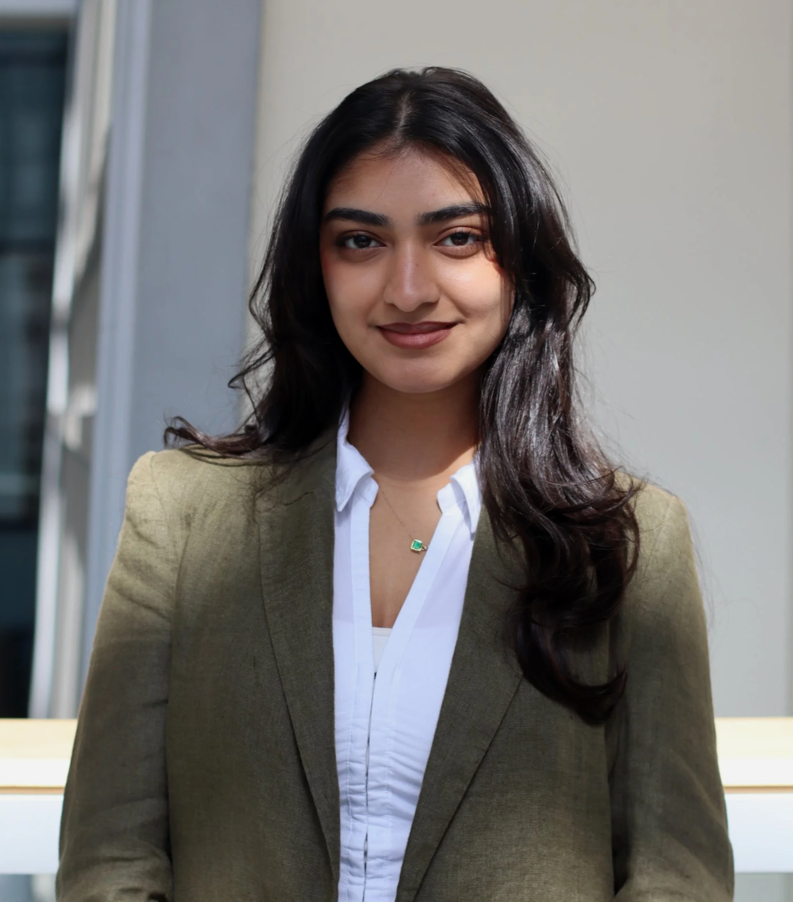 Portrait of a young woman with long dark hair, wearing a white shirt and olive blazer, standing outdoors near a building with windows.