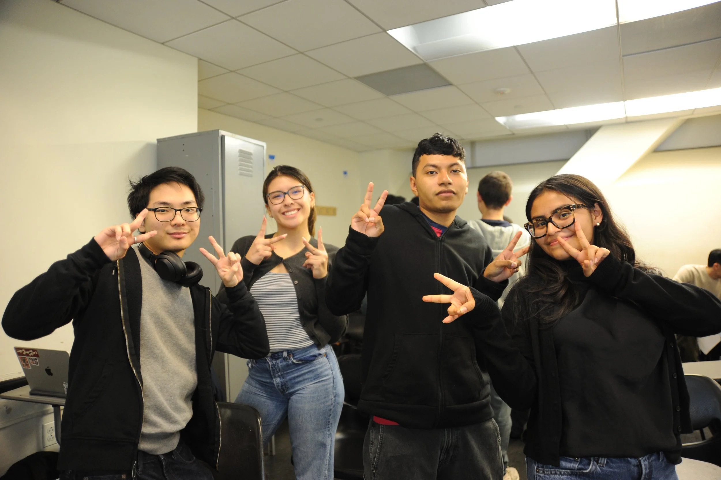 Four friends pose indoors, smiling and making peace signs in a classroom or office.