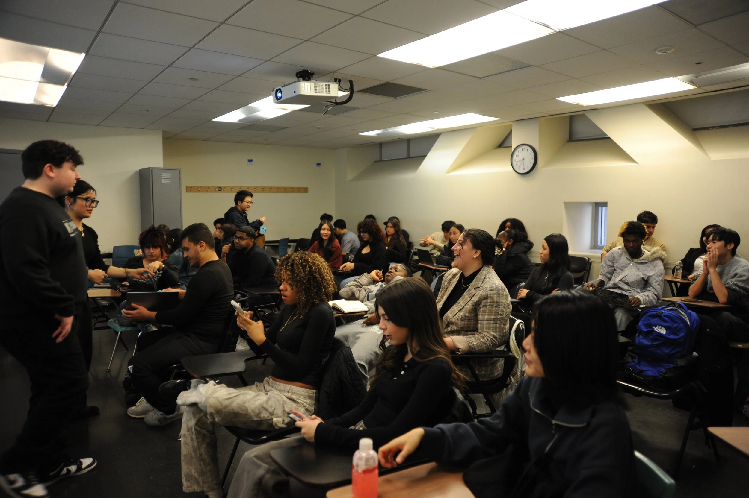 Crowded classroom with students seated at desks beneath a ceiling-mounted projector.