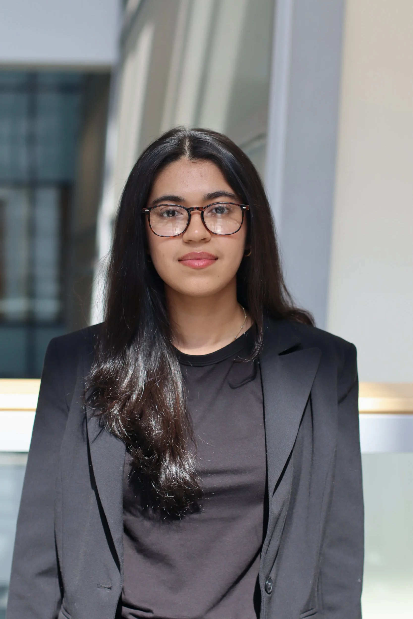 A young woman with long dark hair, glasses, wearing a black blazer and top, standing indoors with modern architecture in the background.
