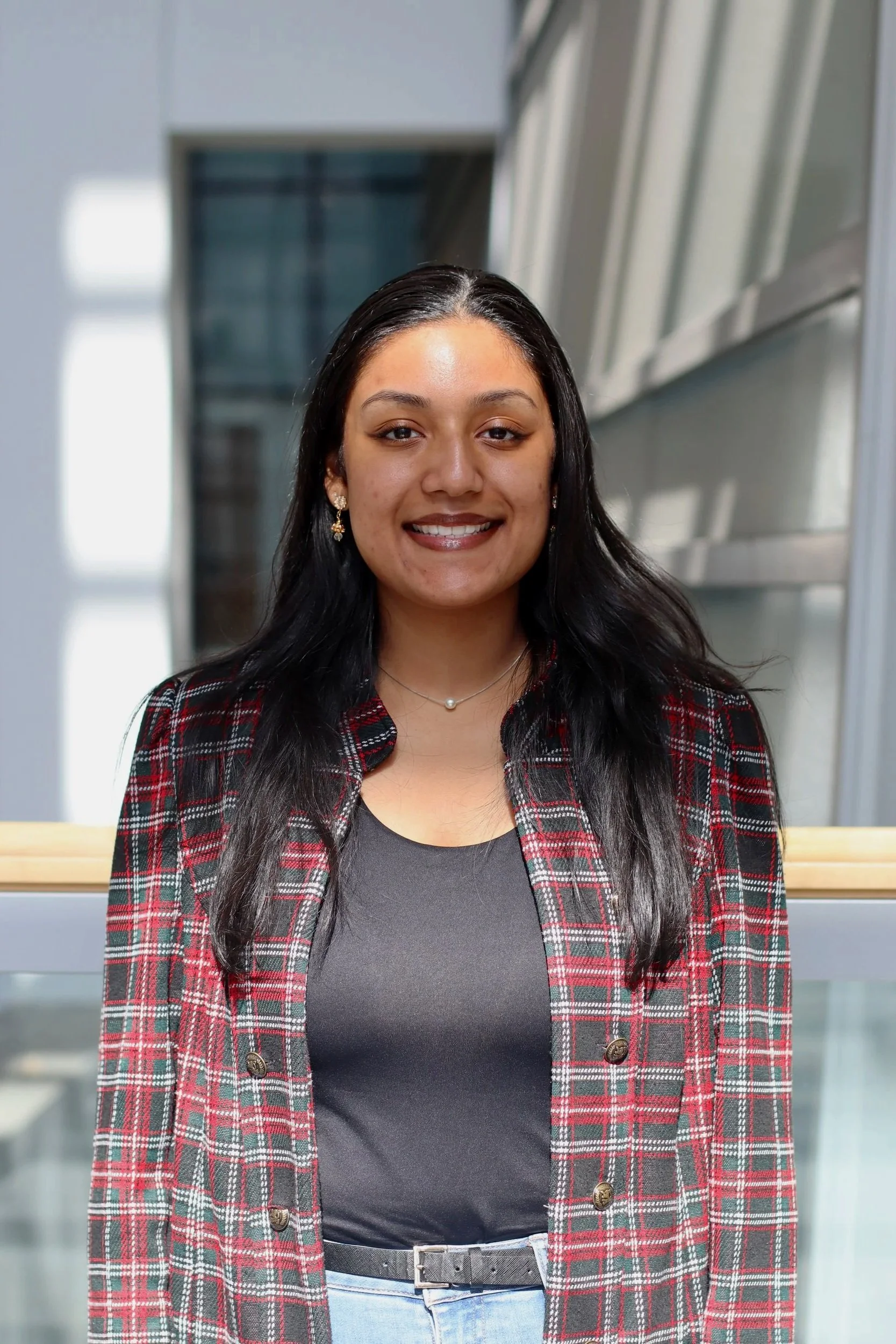 A young woman with long black hair, smiling, wearing a black top, plaid blazer, and light blue jeans, standing indoors near a glass railing with building windows in the background.