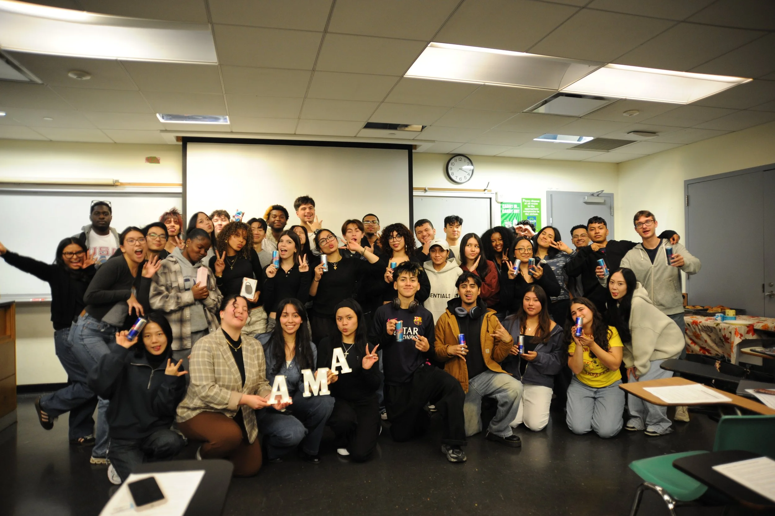 Large diverse group posing for a celebration photo in a classroom, holding drinks and decorative letters.