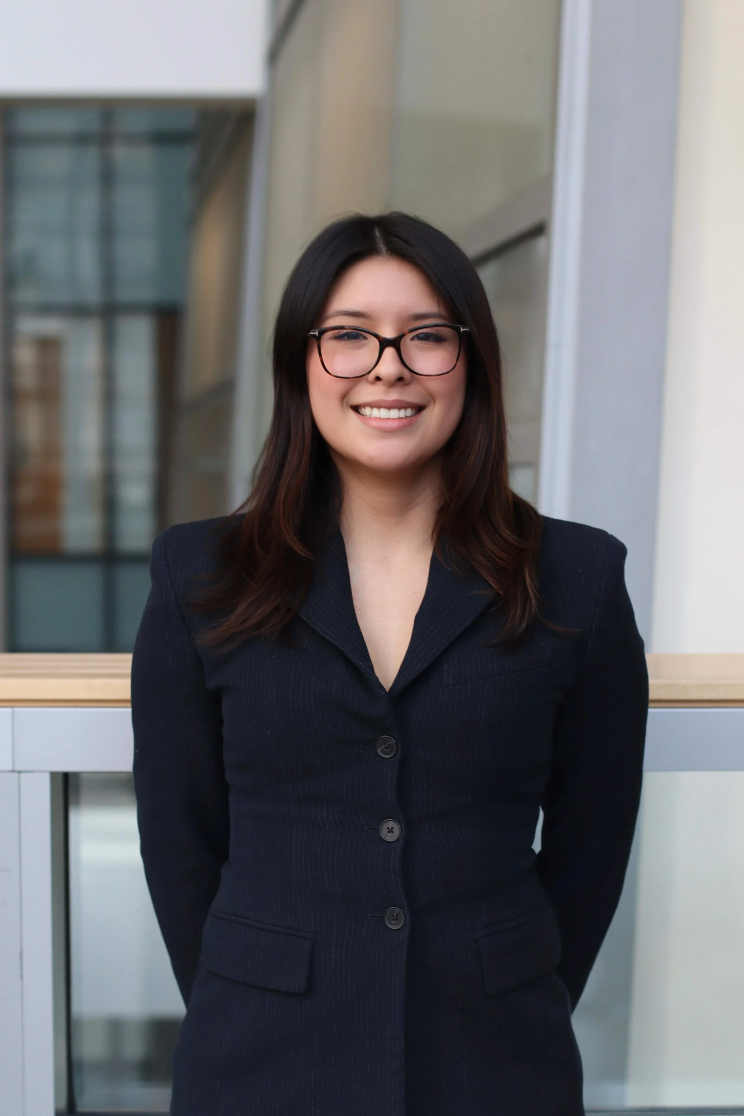 A young woman with shoulder-length dark hair and glasses, smiling in a professional setting, wearing a dark blazer.