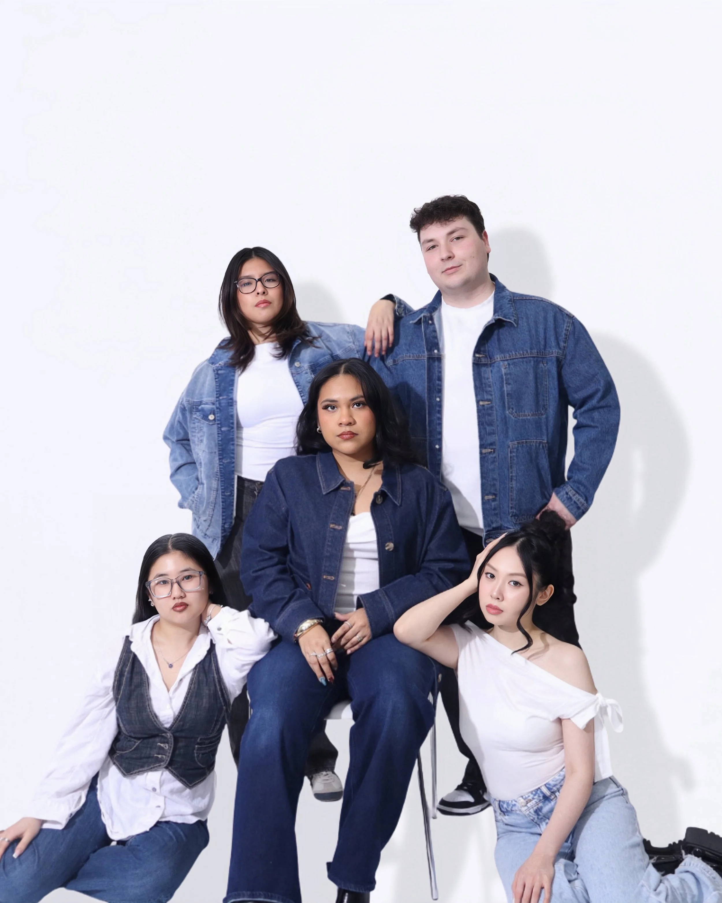 Five young adults posing in front of a white wall, wearing casual denim and white clothing.