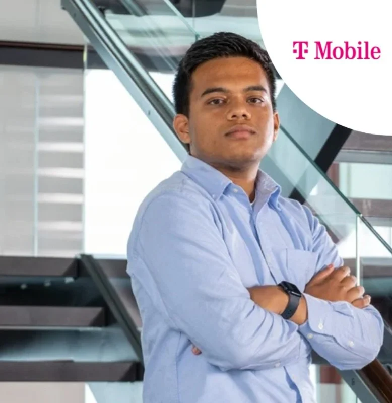A young man in a light blue dress shirt with arms crossed, standing indoors near glass stairs with a T-Mobile logo in the upper right corner.